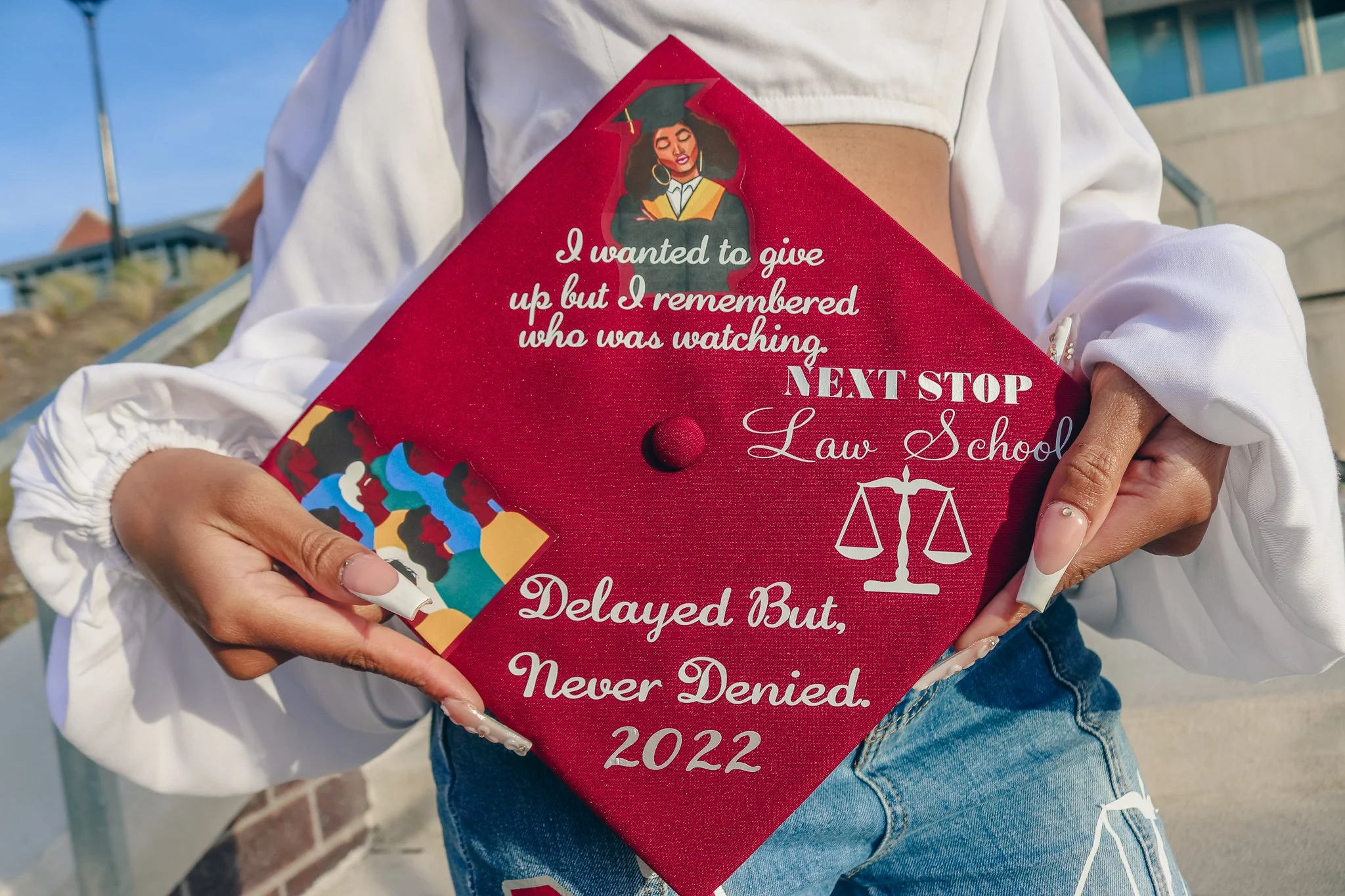 Person holding a red graduation cap with quotation and graphic of a graduate and scales, on a gray shirt and blue jeans, outdoors.