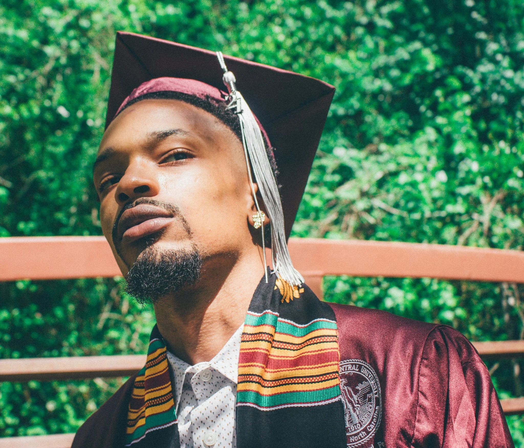 A young man in a graduation cap and gown takes a selfie outdoors, with greenery in the background.