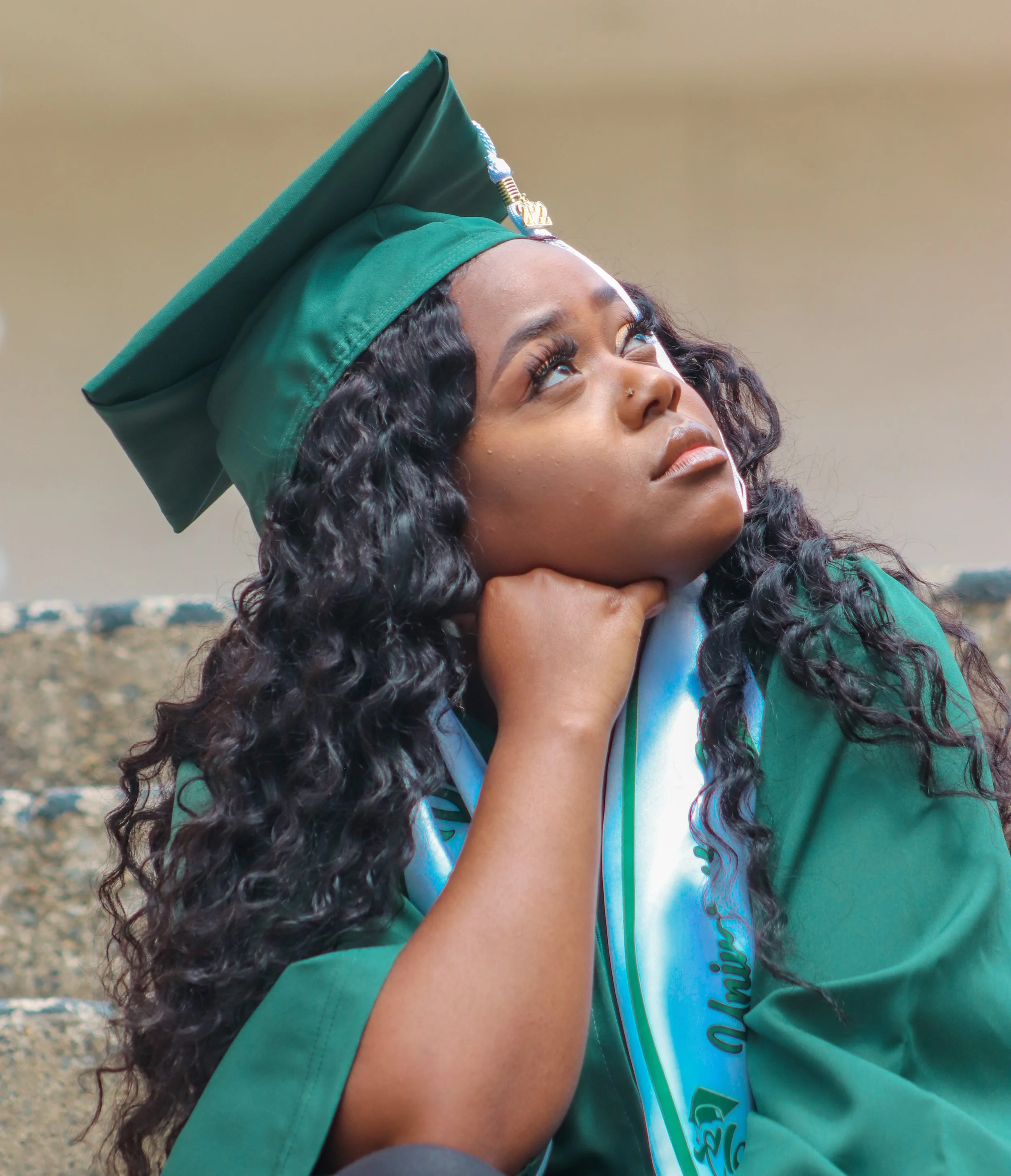 A young woman wearing a green graduation cap and gown, sitting with her chin resting on her hand, looking upward thoughtfully.