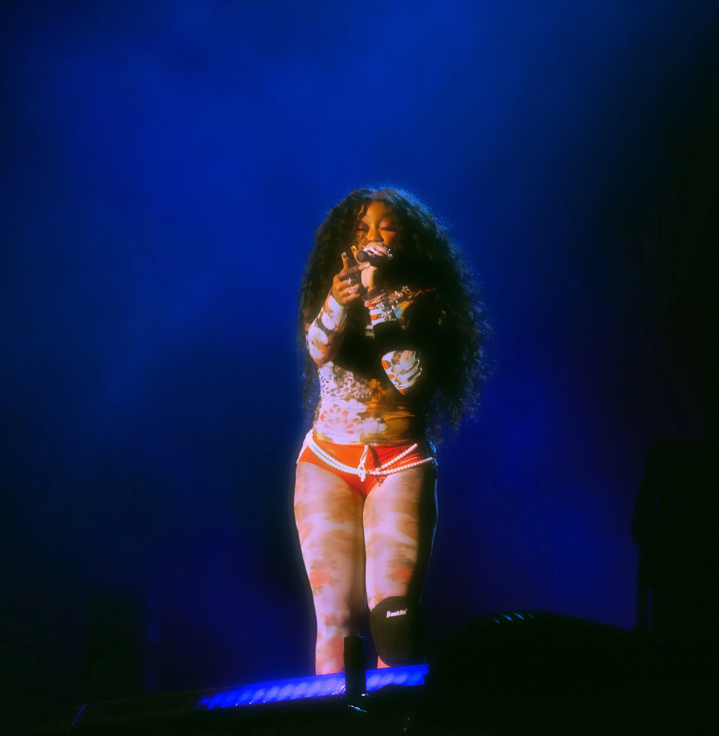 A woman with long curly hair performing on stage under blue lighting, wearing a floral top, red shorts, and a black knee brace, holding a microphone.