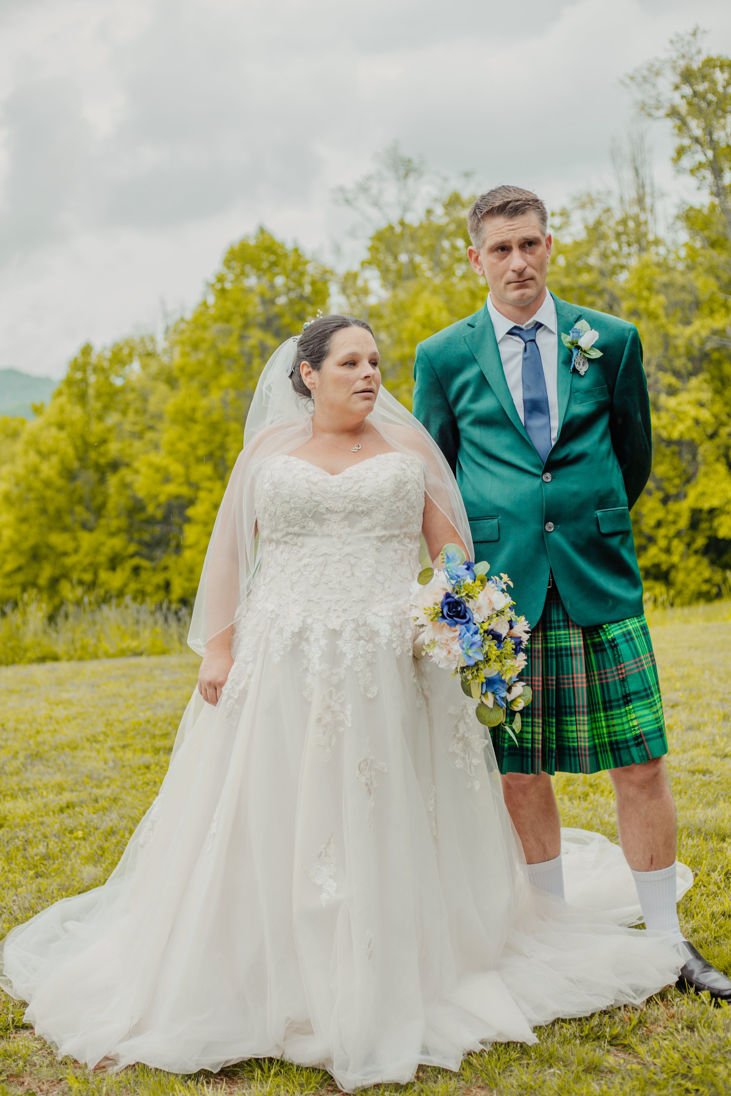 A bride and groom standing outdoors in a field, with the bride in a white wedding dress and the groom in a green tartan kilt and jacket, both looking somber.
