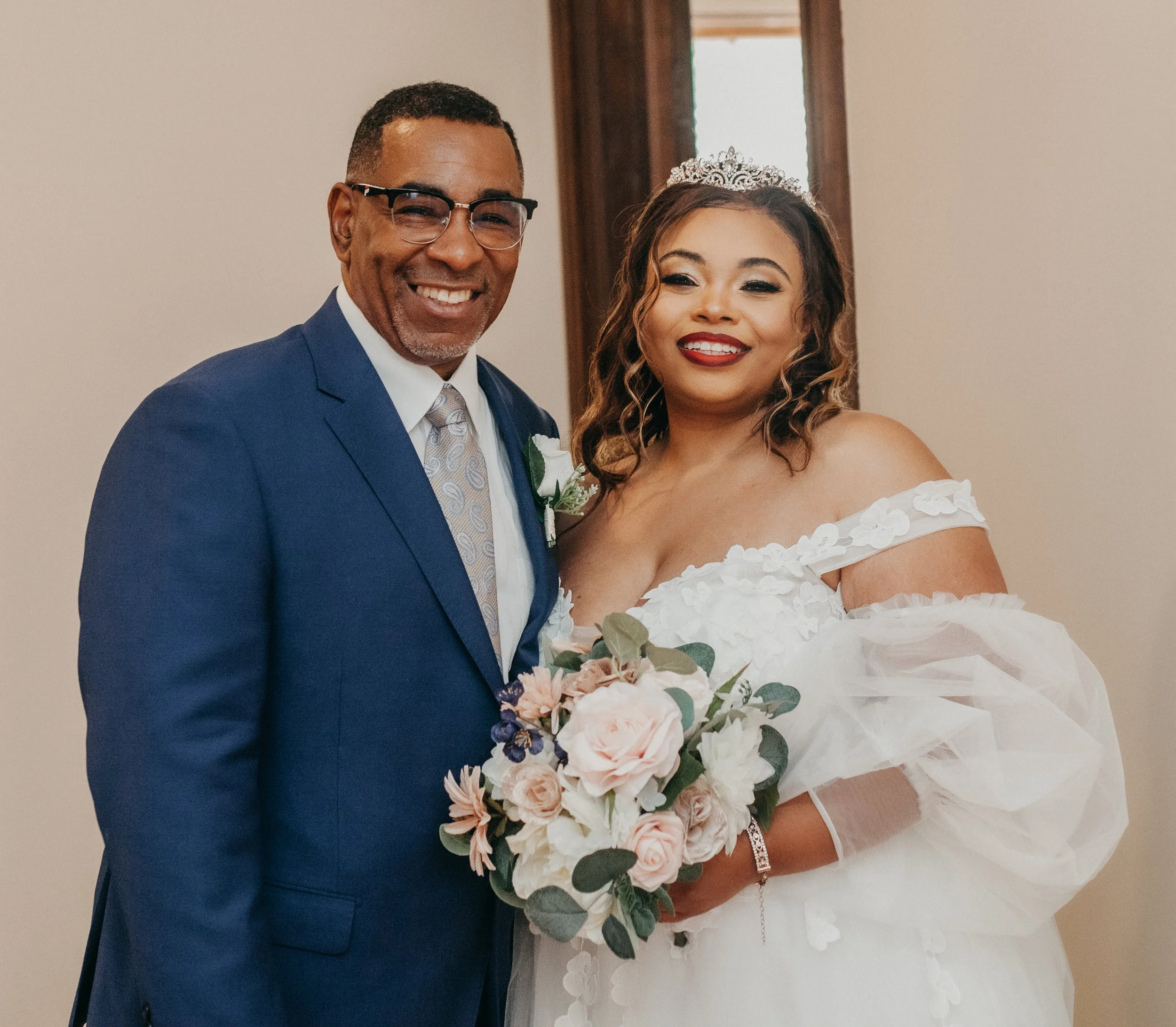 A smiling bride in a white off-shoulder wedding dress holding a bouquet of pink and white roses, standing next to a smiling man in a blue suit with glasses, in an indoor setting.