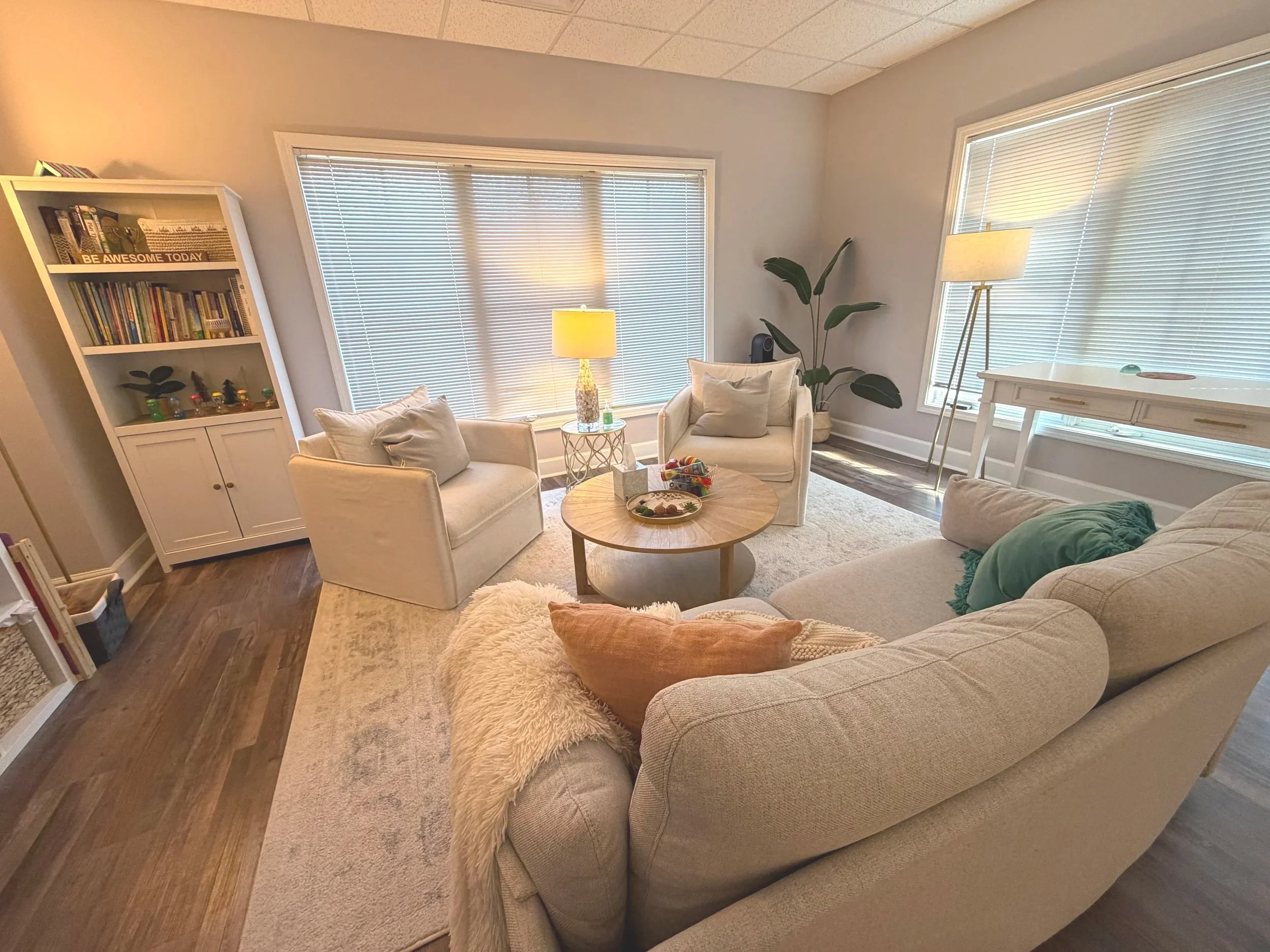 A cozy living room with a beige sofa, two matching armchairs, a round coffee table, a white bookshelf, two large windows with blinds, a floor lamp, a side table with a lamp, and decor including pillows, plants, and books.