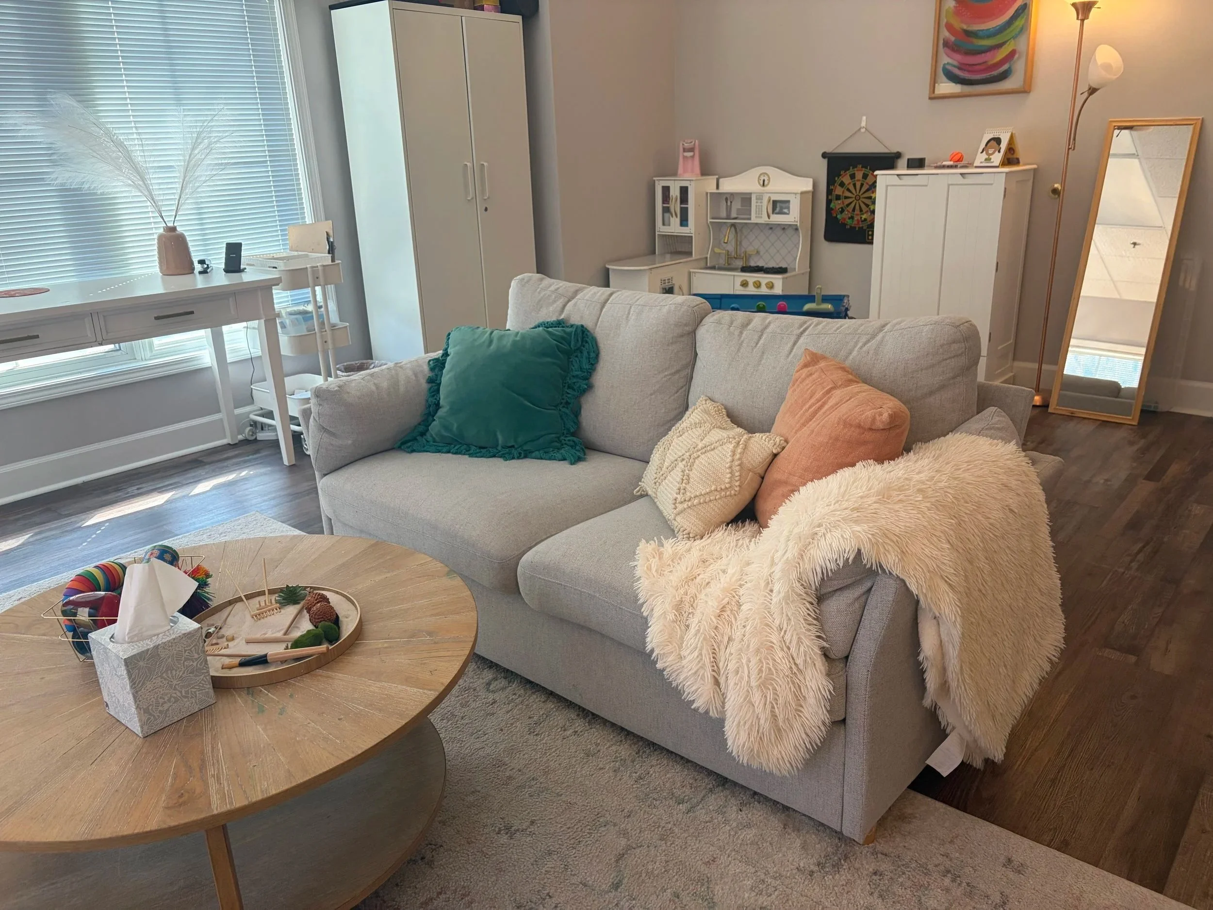A cozy living room with a light gray sofa decorated with green, peach, and cream pillows, a fluffy cream blanket, a wooden coffee table with colorful yarn, a tissue box, and jewelry display. In the background, a white cabinet, a dollhouse, a mirror, 