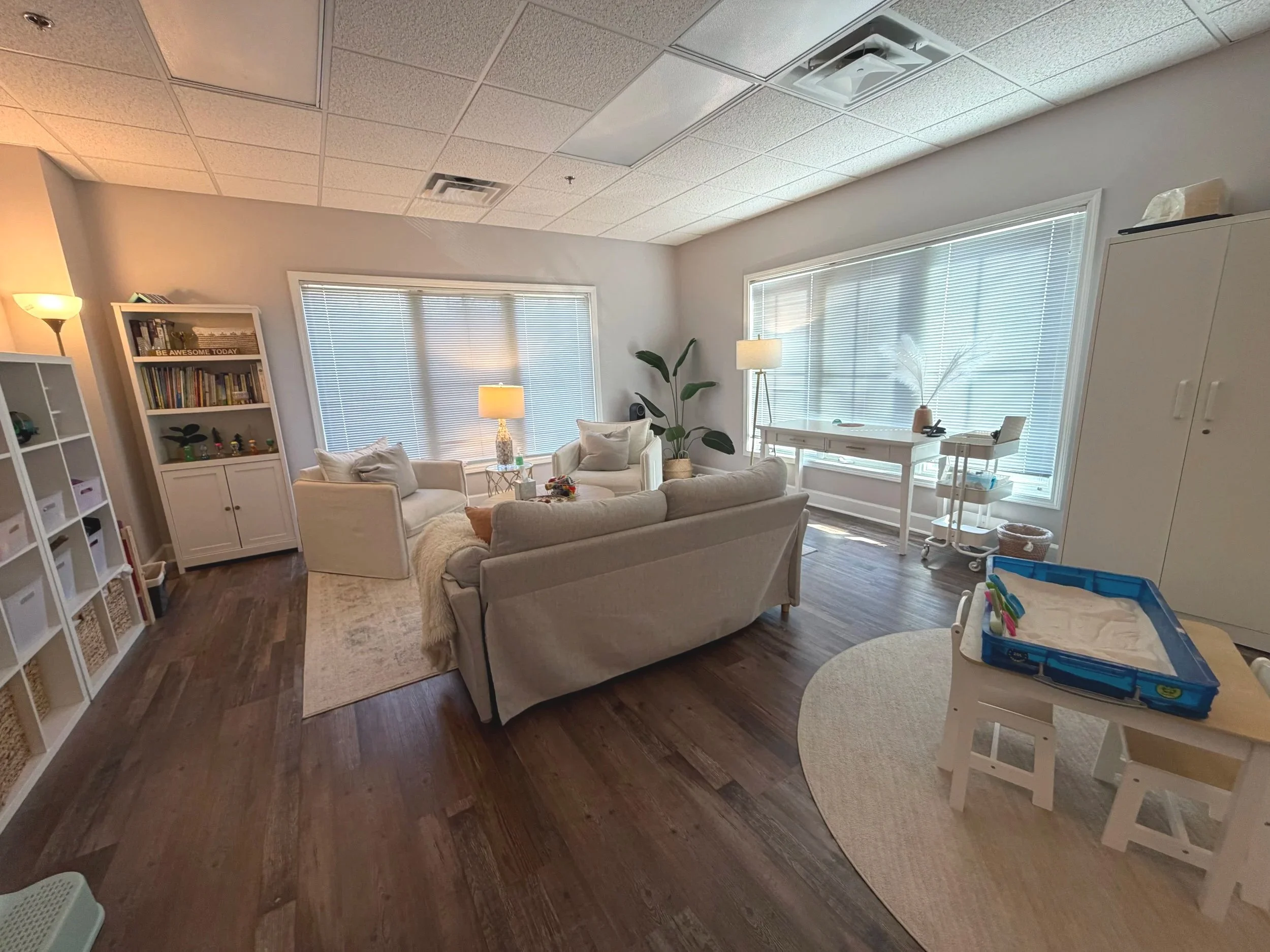 Bright living room with white couches, wooden floors, large windows with blinds, bookshelf, and potted plant.