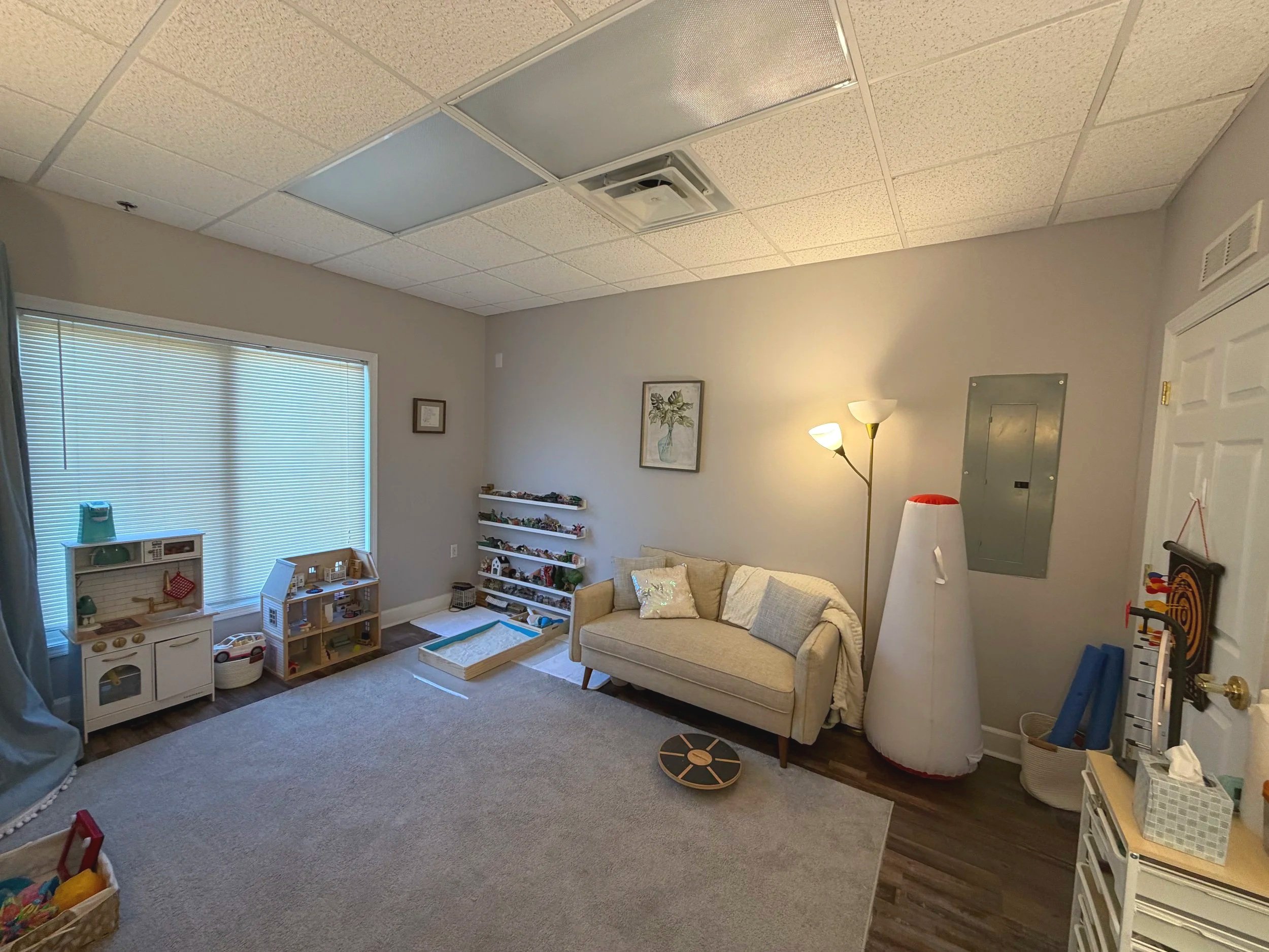 A cozy children's playroom with a large window covered by closed blinds, a beige sofa with decorative pillows, a tall floor lamp, a white vertical lamp, wall-mounted shelves with toys, and various children's toys and furniture.