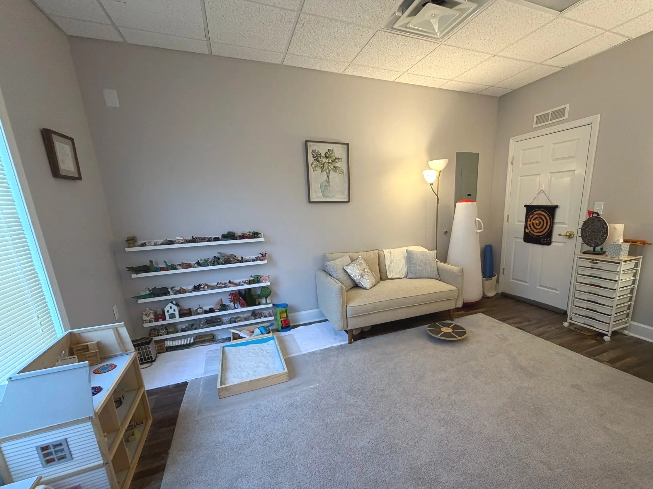 Living room with shelves of toys, a cream-colored sofa with pillows, a lamp, a small table, a dartboard, and a white door.