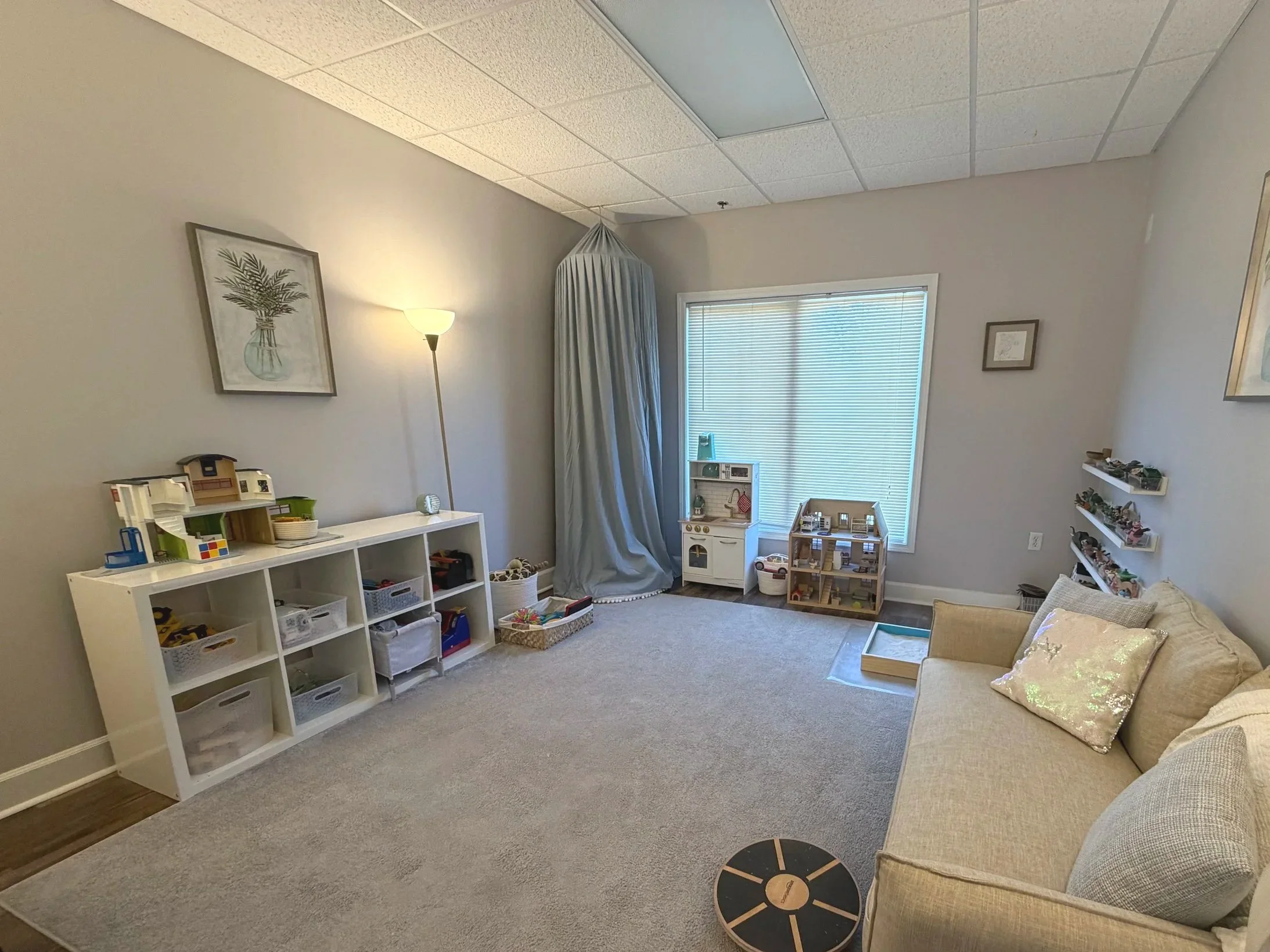 A living room with a beige sofa, a window with blinds, shelves with toys, a lamp, and a framed picture on the wall.