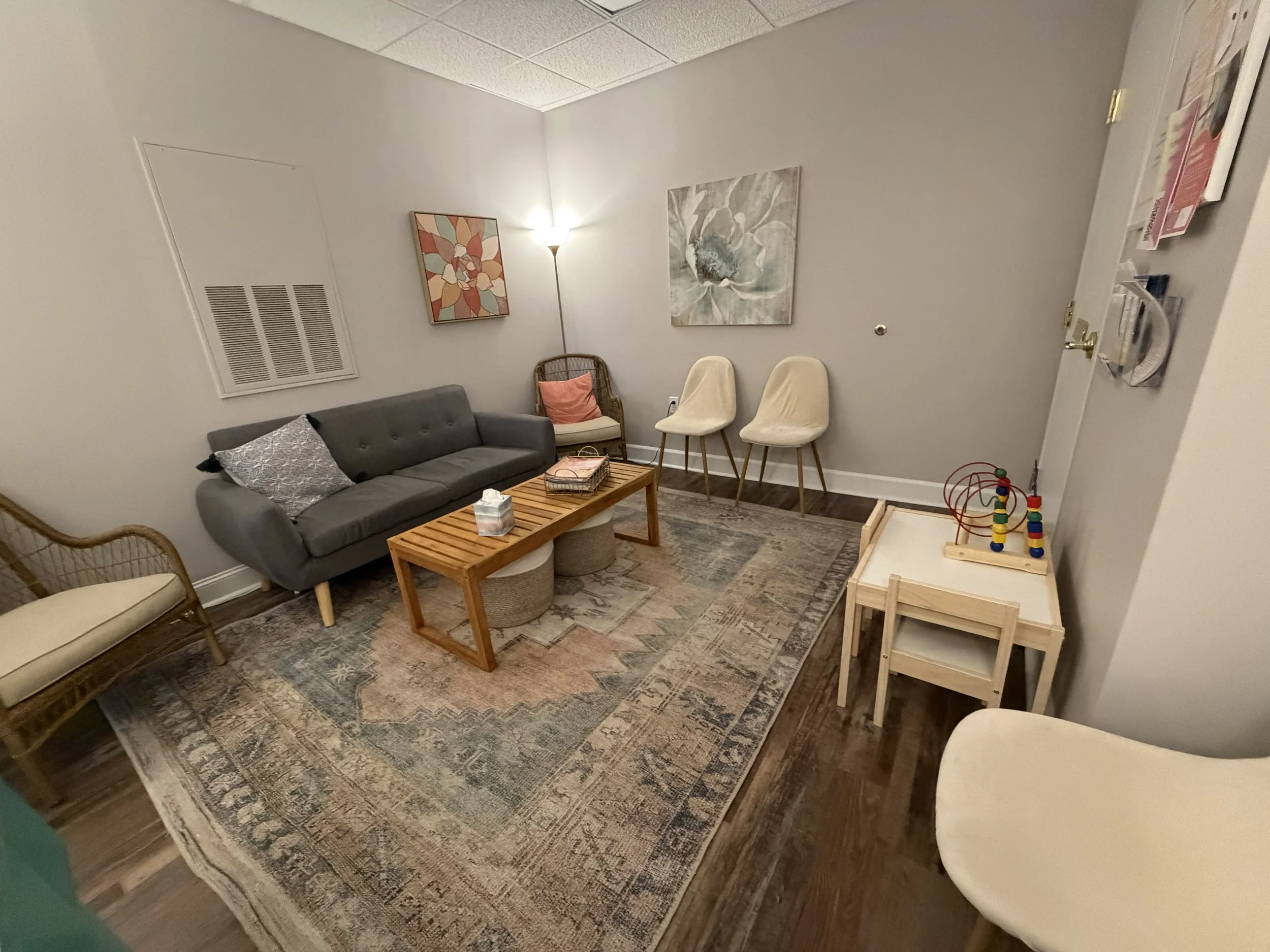Living room with a gray couch, wooden coffee table, white chairs, a wicker chair, and a children's toy table with bead mazes, decorated with artwork and a floor lamp.