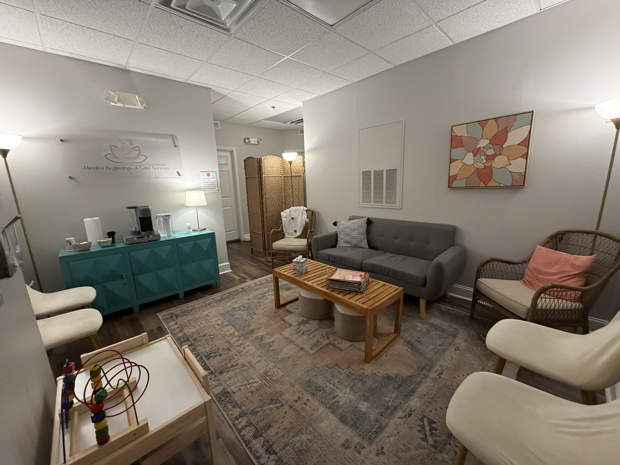 A cozy waiting room with a gray couch, patterned armchair, and a coffee table on a rug. There are white and wicker chairs, a teal cabinet with a coffee maker, and artwork on the walls. The room is softly lit by floor lamps and a table lamp.