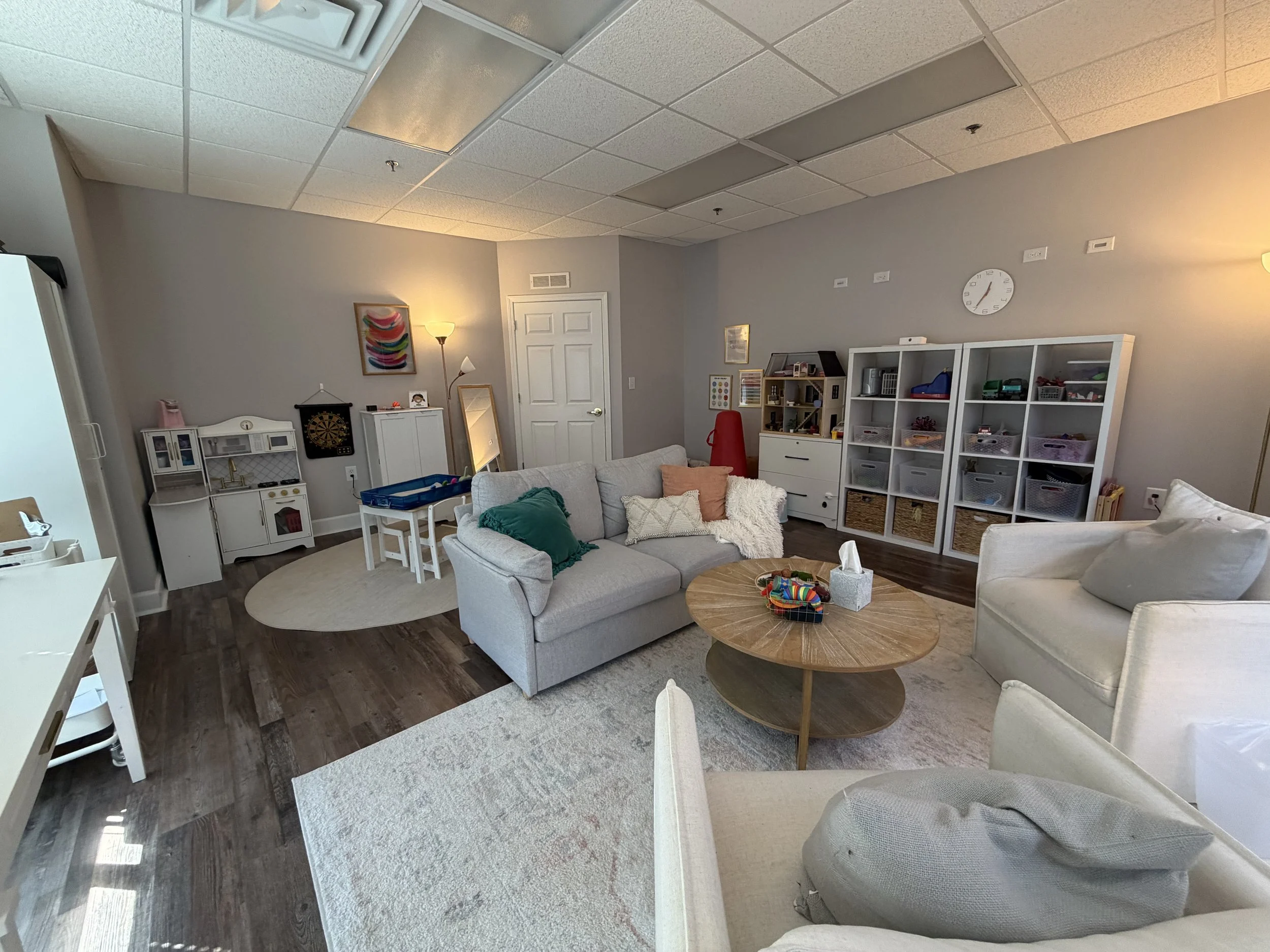 Living room with white sofa, a white armchair, a wooden coffee table with tissue box and toys, and a play kitchen set in the corner. There is a shelving unit with storage baskets, and a clock on the wall. A lamp and artwork are also visible.