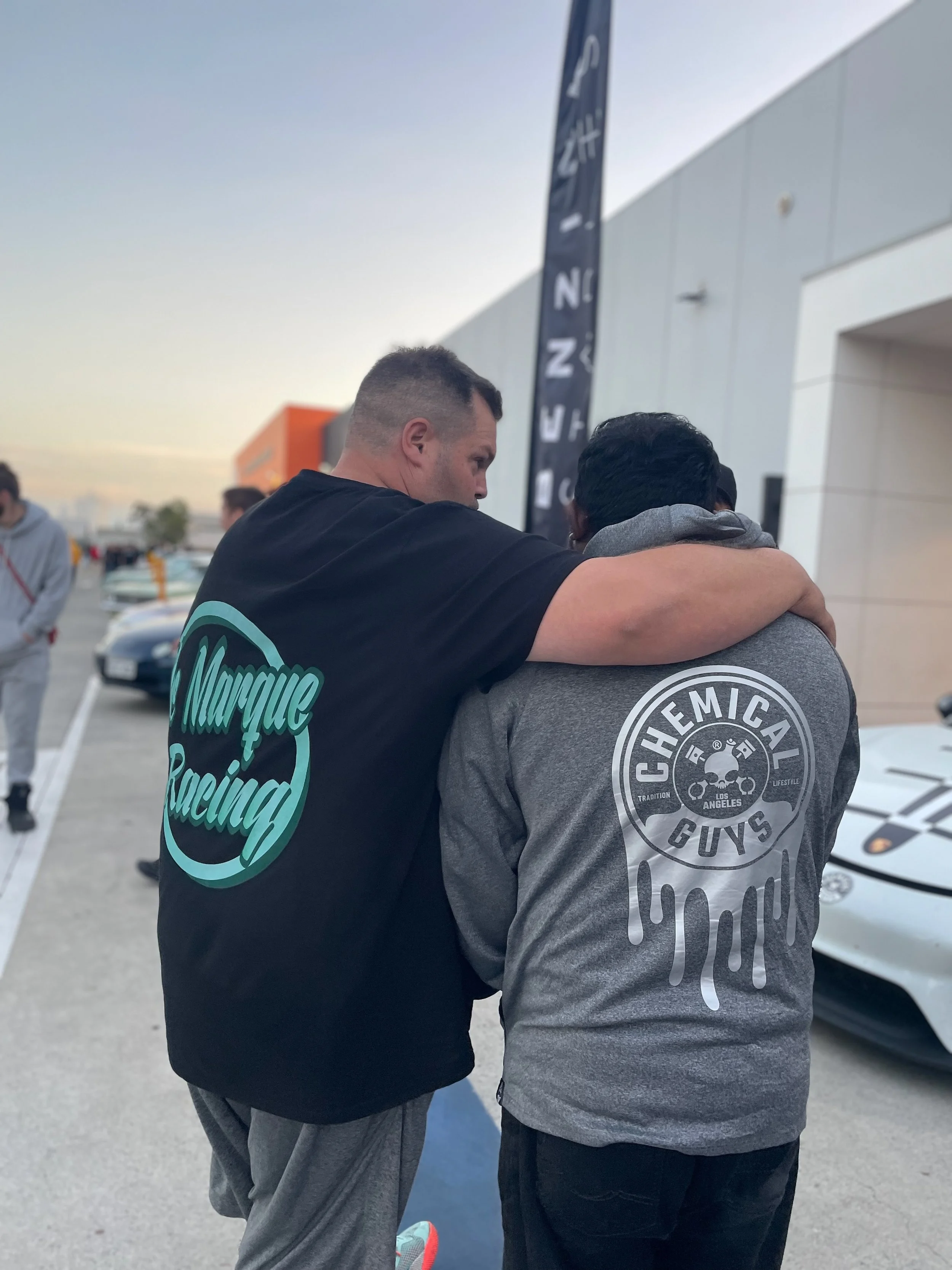Two people standing together, one wearing a 'Marque Racing' T-shirt and the other wearing a 'Chemical Guys' hoodie. They have their arms around each other, with cars and a building in the background.