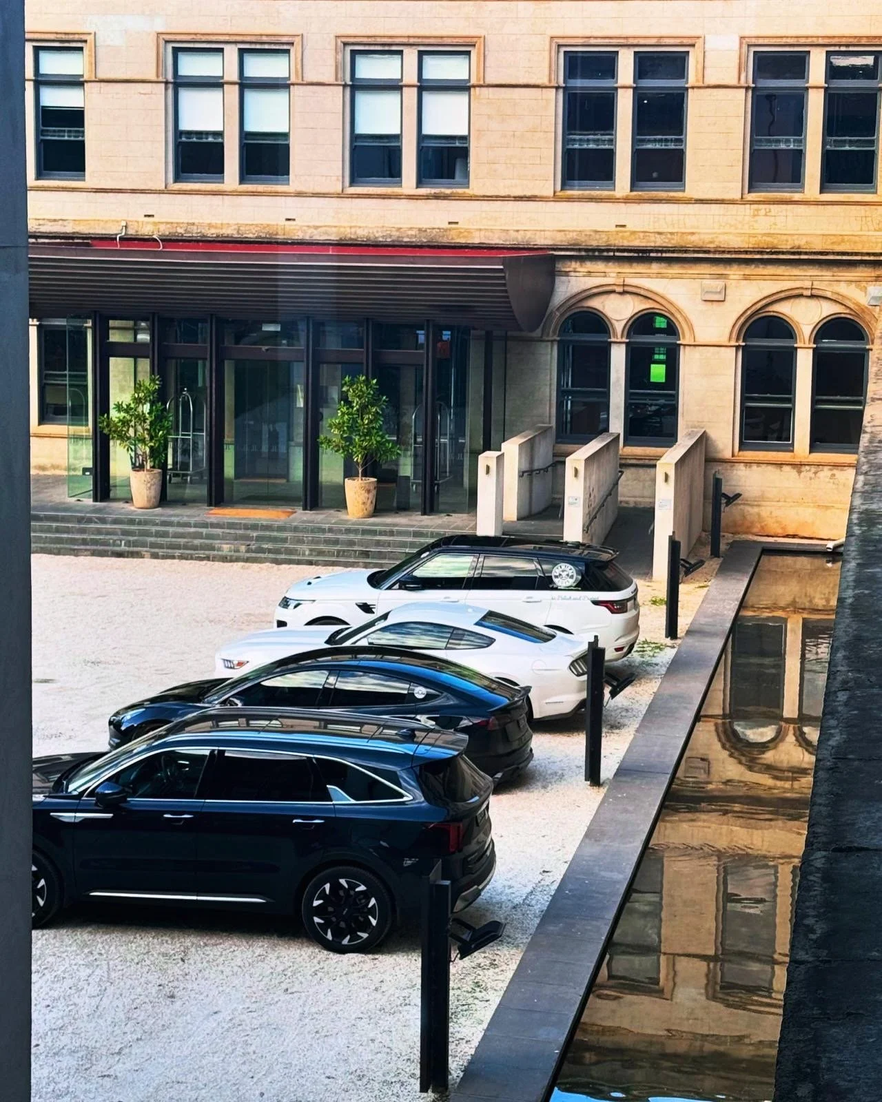 Parking lot with four parked vehicles in front of a building with large windows and exterior steps. The scene includes a reflection of the building in a puddle or water feature.