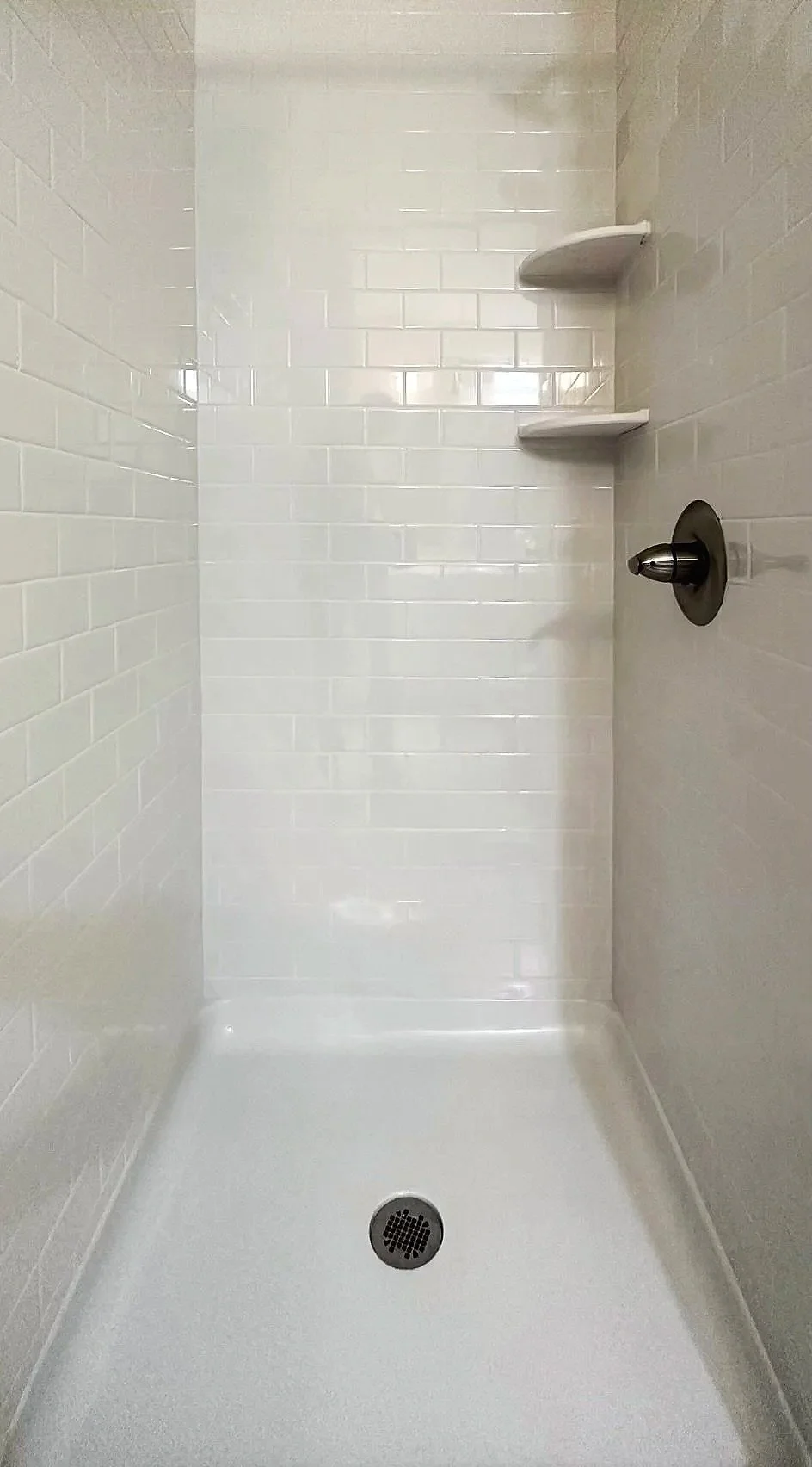 Empty shower stall with white subway tile walls, a brushed nickel shower fixture, and built-in corner shelves.