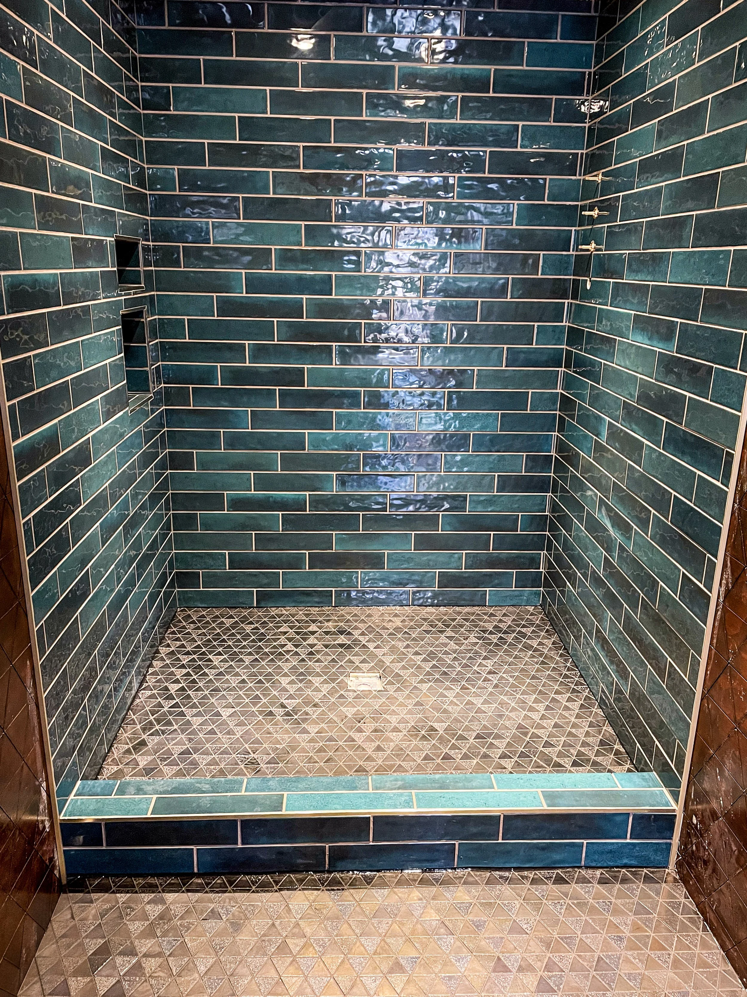 An empty shower with teal subway tiles on the walls, small brown mosaic tiles on the floor, and built-in wall niches.