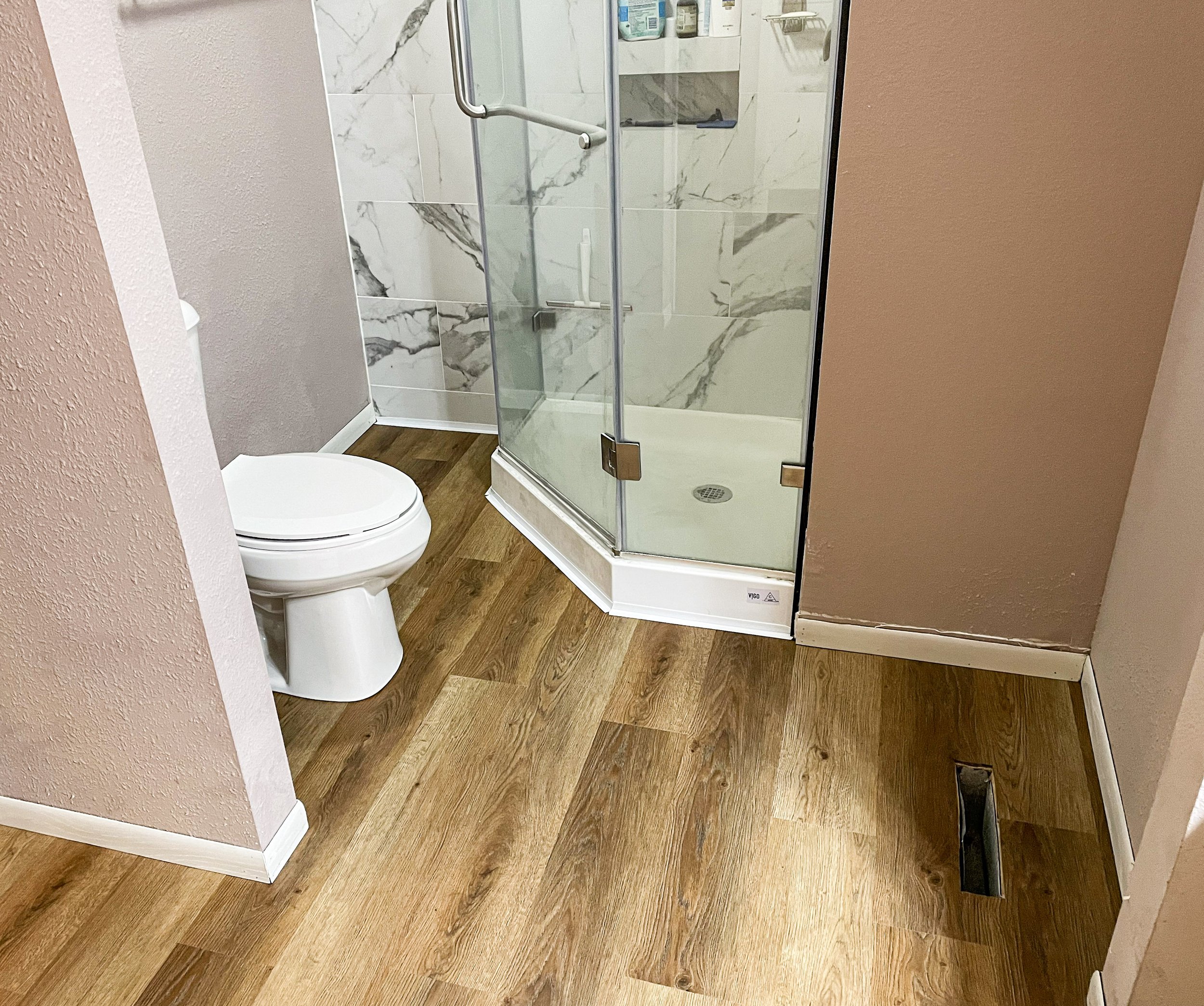 A bathroom with a wood-look LVP floor, a white toilet, and a glass shower stall with marble tiles.