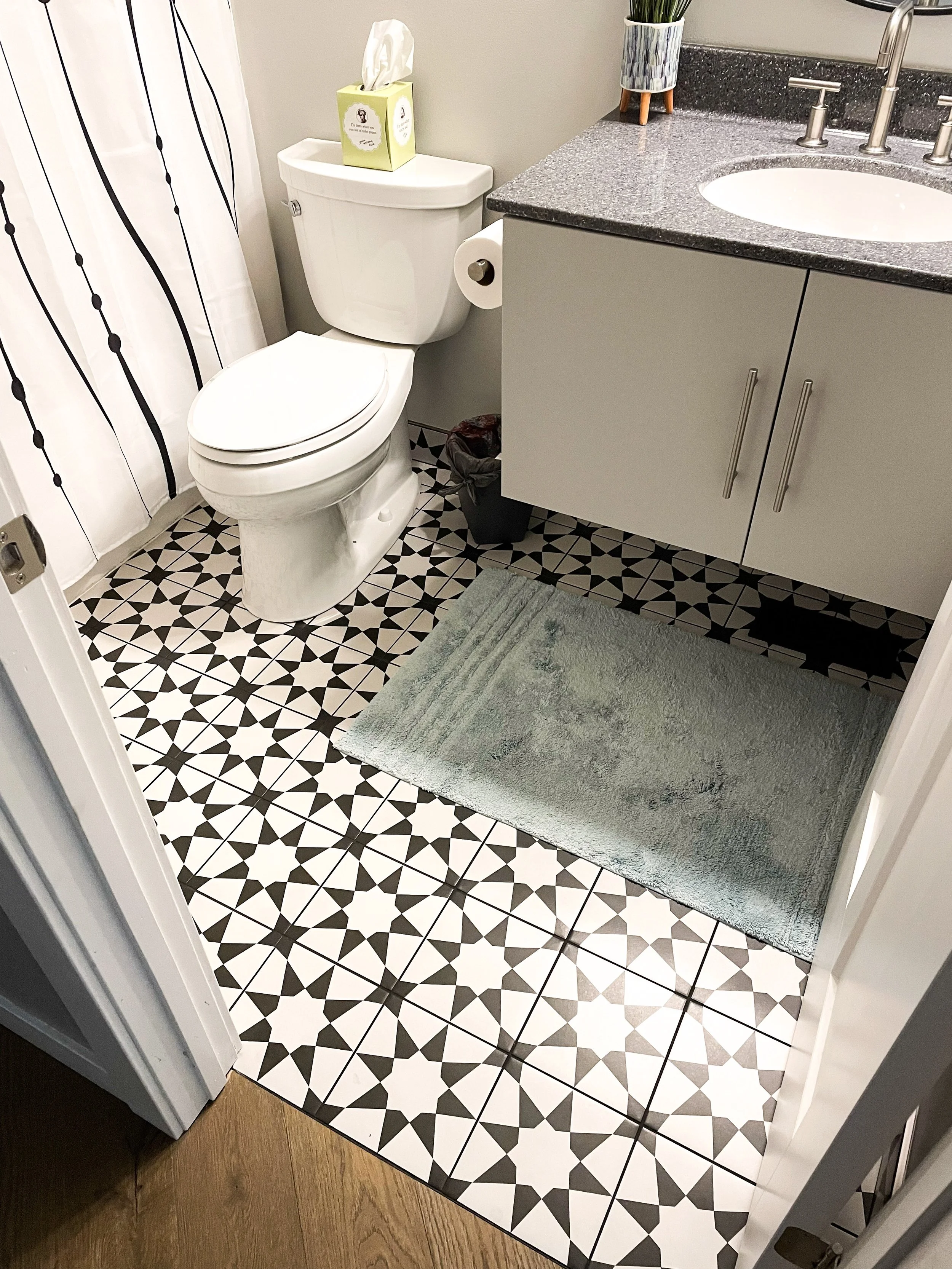 A small bathroom with black and white geometric patterned floor tiles, a white toilet with a box of tissues on top, a gray vanity with a speckled gray countertop, a sink with silver faucets, a small potted plant, a gray bath mat, and a shower curtain
