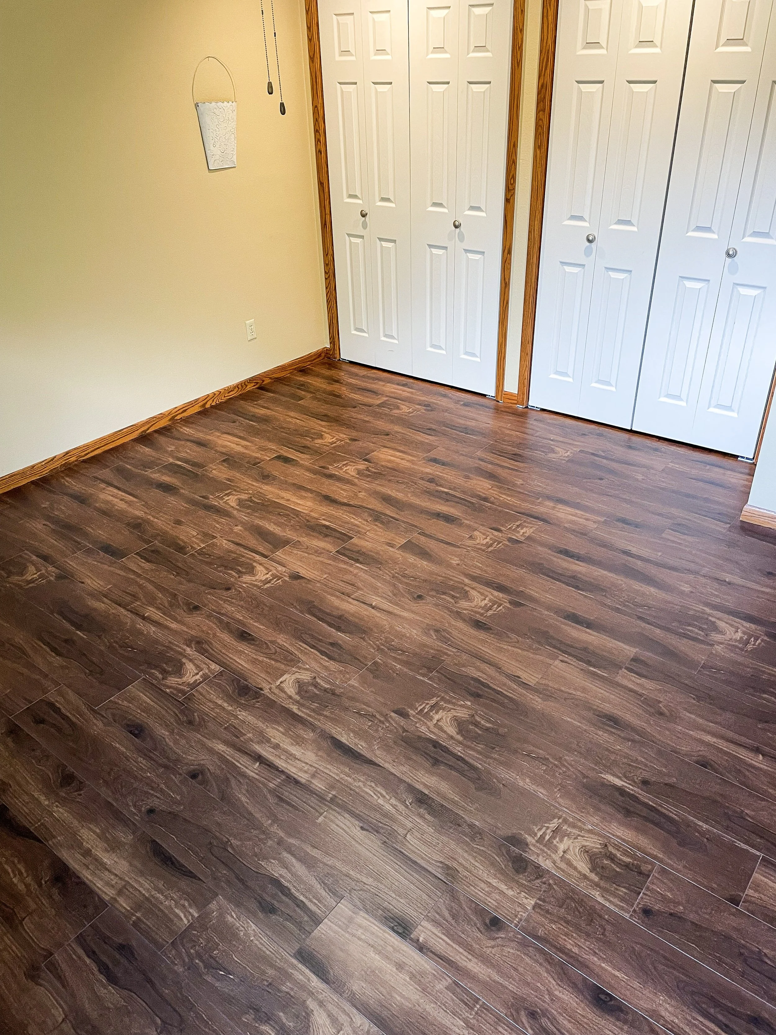 Empty room with wood laminate flooring, beige walls, a wall hanging, two closed white closet doors, and a door frame.