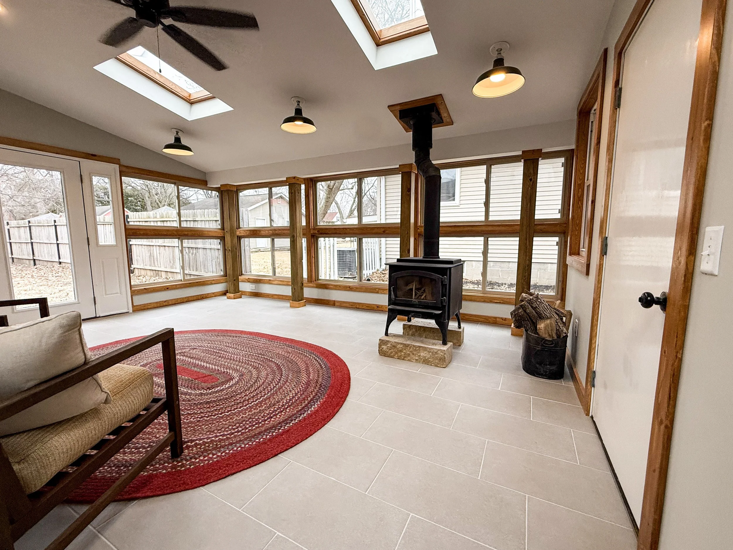Sunroom with large windows, wood trim, a black wood stove, and a red circular rug on gray tile flooring.