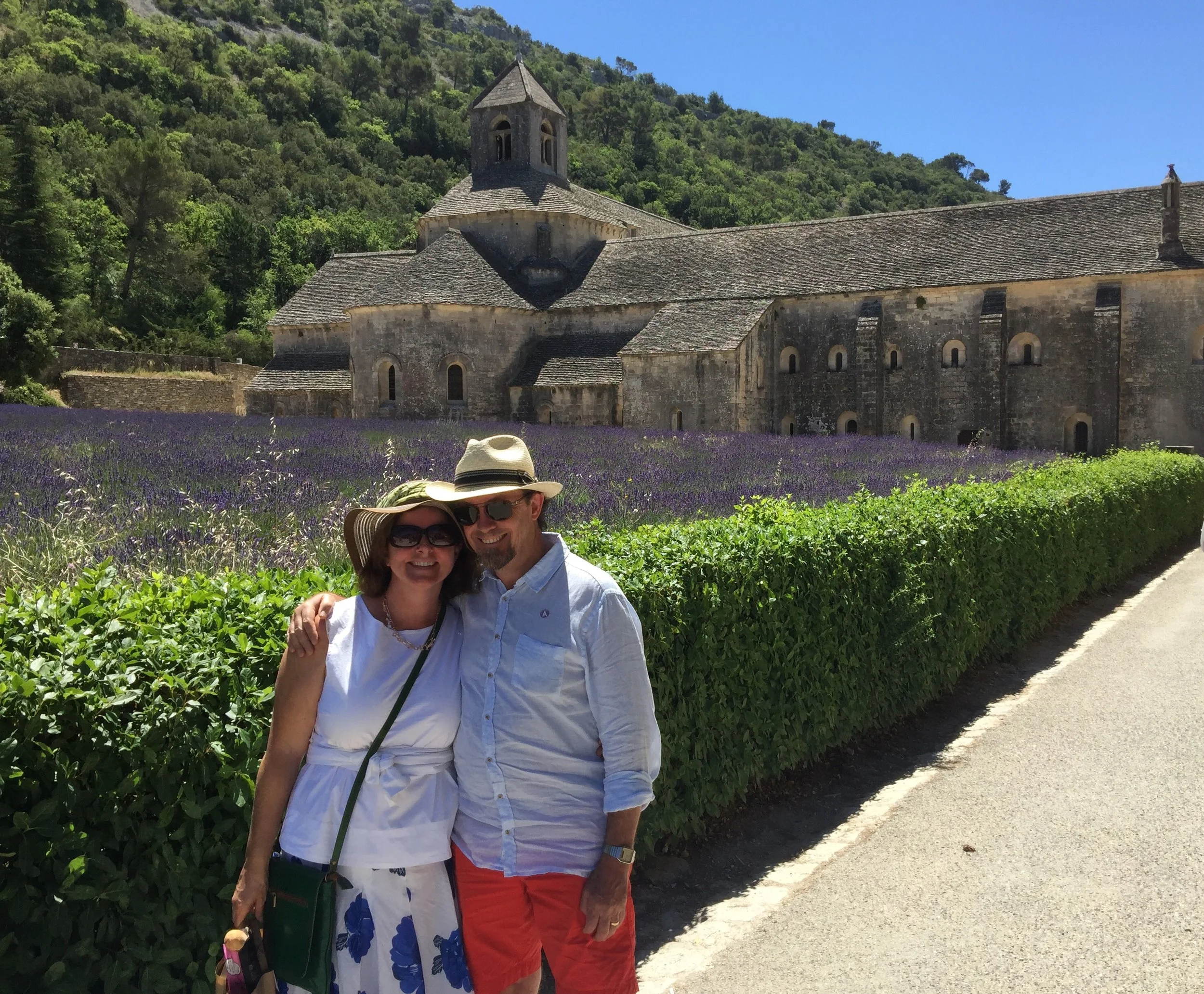 A couple standing outdoors in front of a field of lavender with an old stone church and green hillside in the background, blue sky.