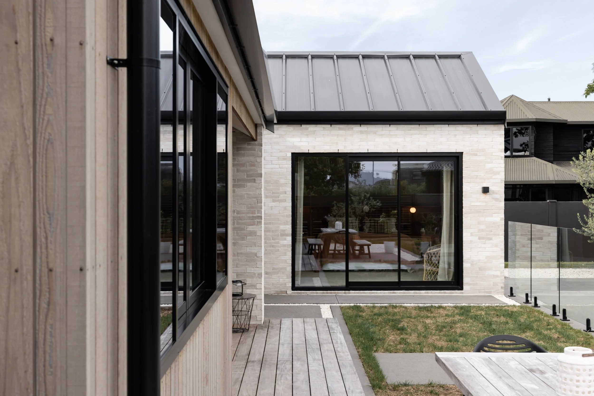 Exterior view of a modern house with a wooden deck, large glass windows, a white brick wall, and a metal roof, with a lawn and small garden area.