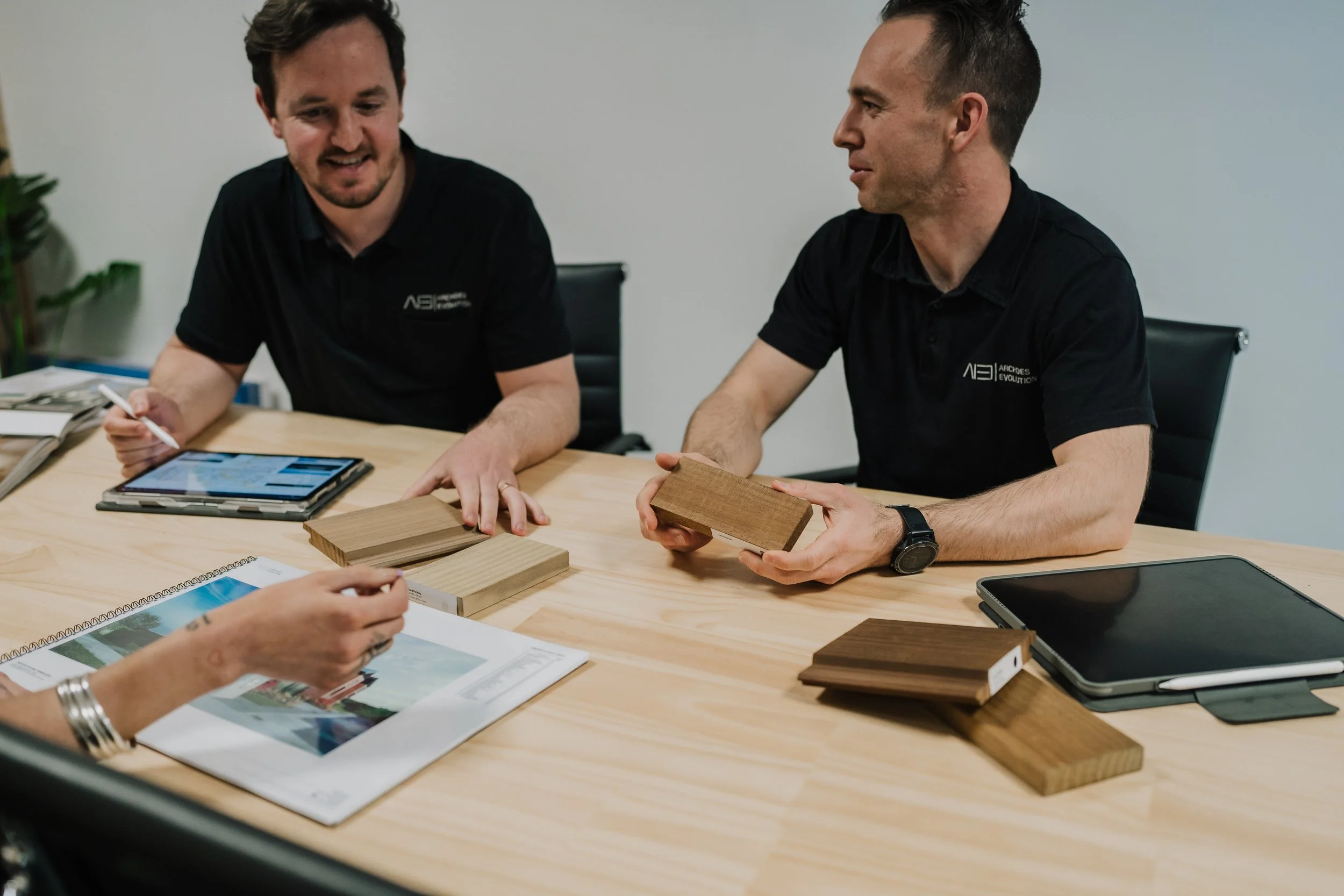 Two men sitting at a conference table discussing wood samples, with a woman showing a brochure and digital tablet in a meeting room.