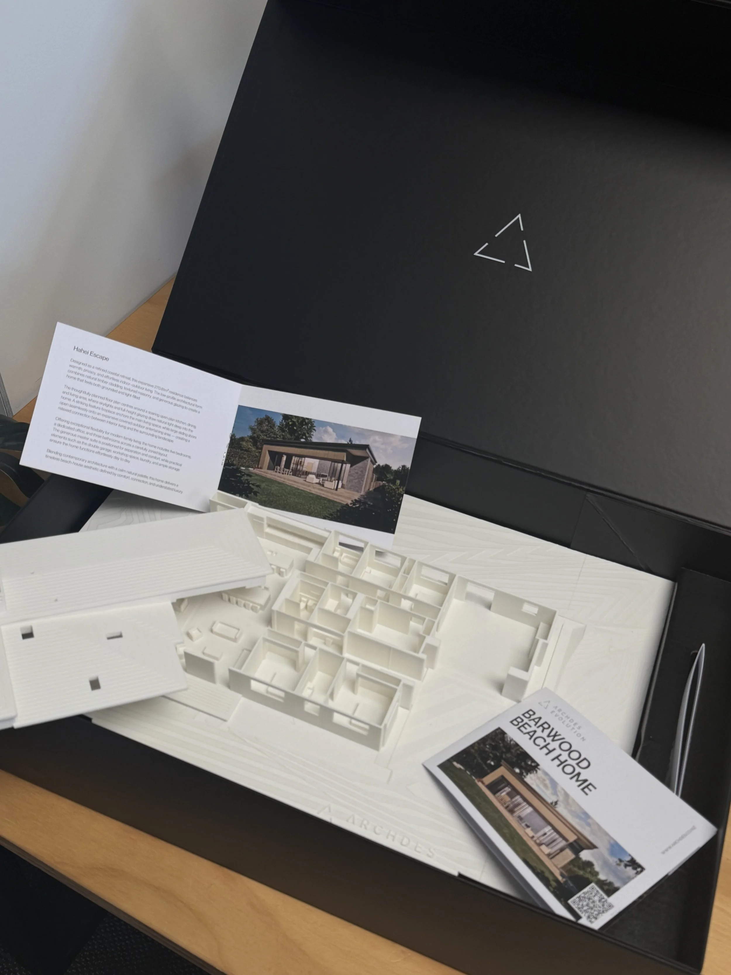 Architectural models, brochures, and printed drawings of a modern home design on a table, with a black presentation board in the background.