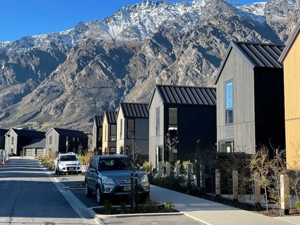 Modern houses with gray and yellow panels on a street in a mountainous area with snow-capped peaks in the background. Several parked cars are visible along the street.