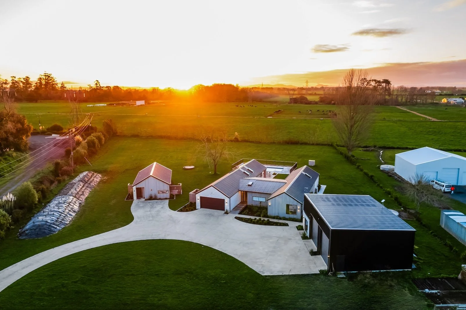 A modern house with multiple sections and solar panels on the roof, situated in a large grassy yard with a curved concrete driveway, late sunset sunlight, and fields in the background.