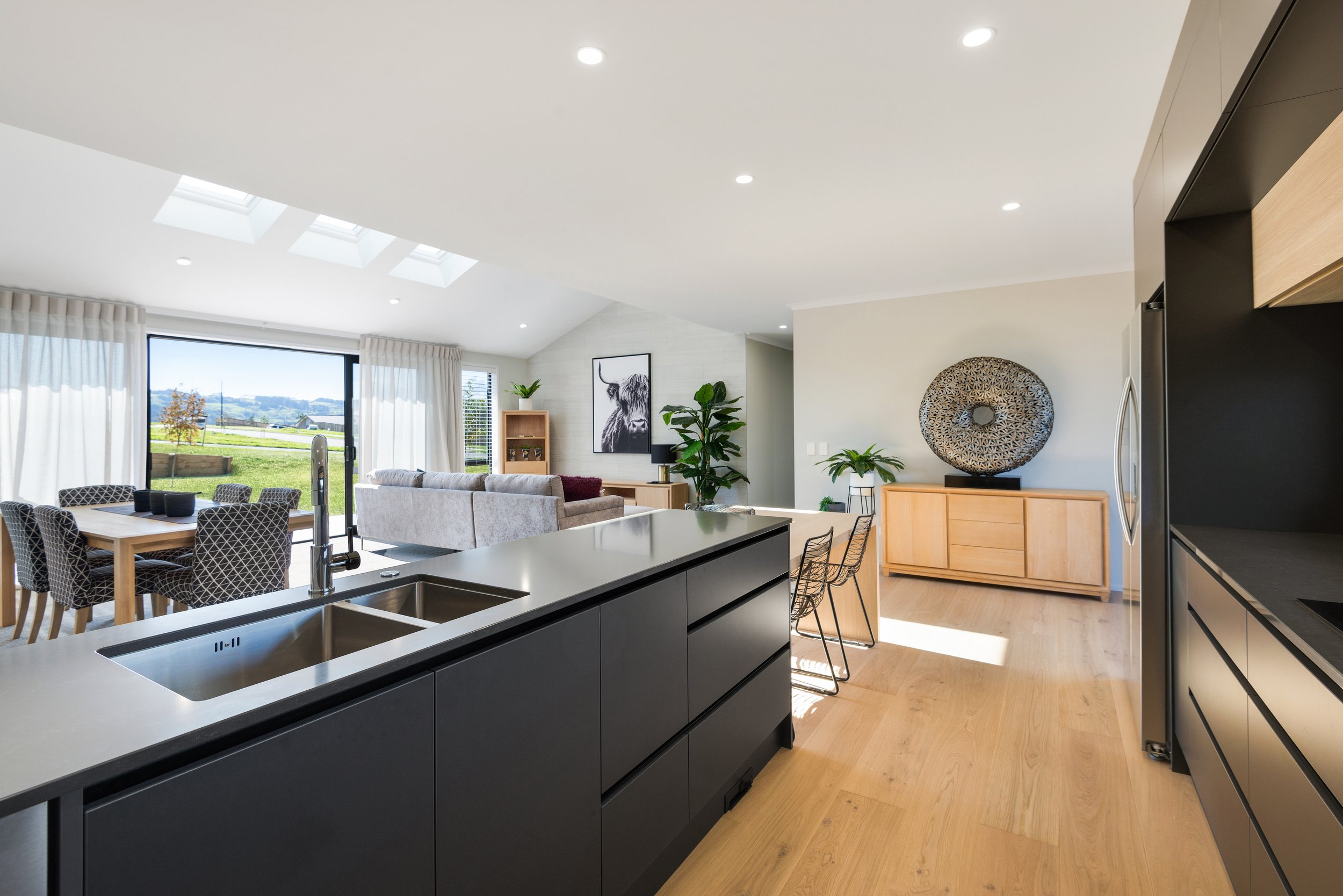 Open-concept living room and kitchen with large windows, white curtains, indoor plants, black and gray furniture, and decorative wall art.
