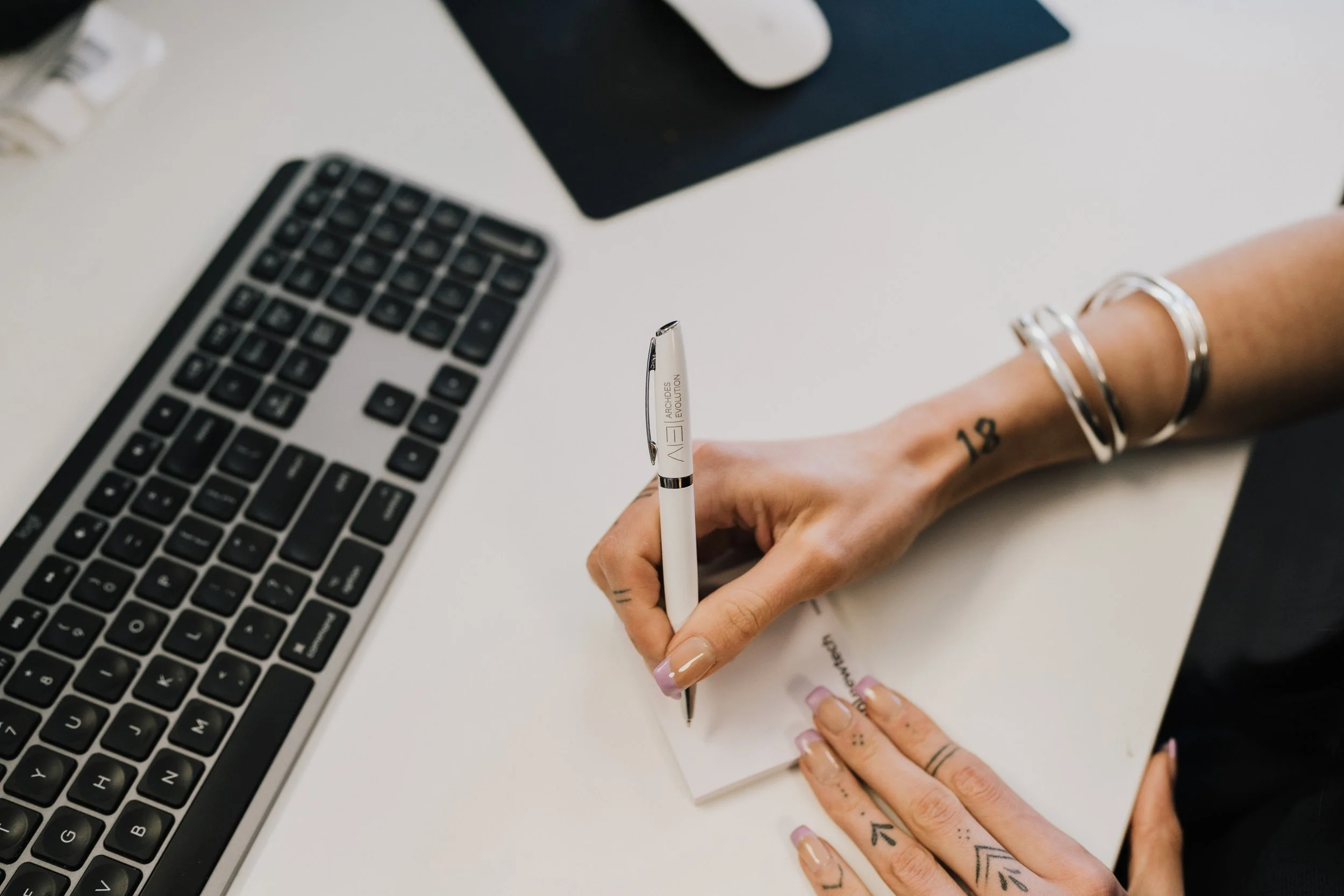 A person writing on a notepad with a black pen, nearby a computer keyboard on a white desk.