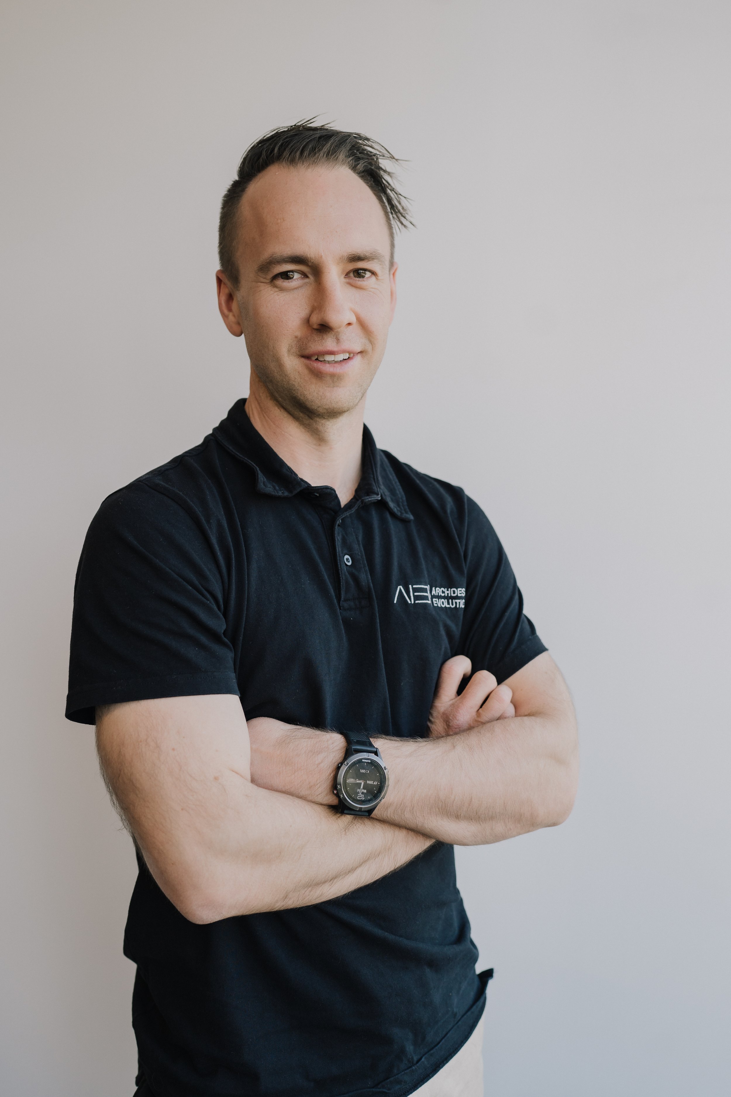A man with short dark hair, wearing a black polo shirt with a logo, has arms crossed, standing against a plain wall.