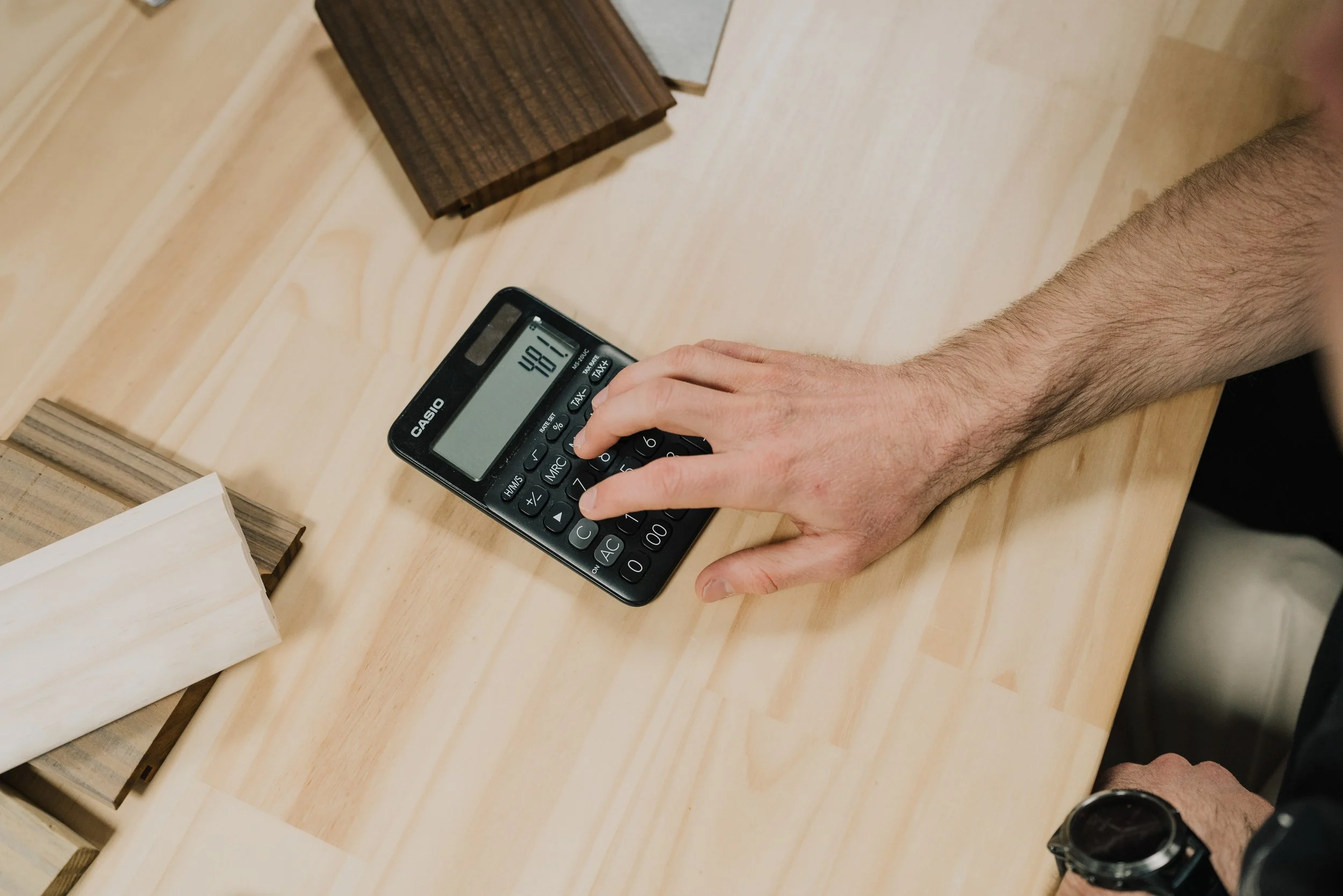 A person's hand using a calculator on a wooden table near small wooden blocks and a white tissue.