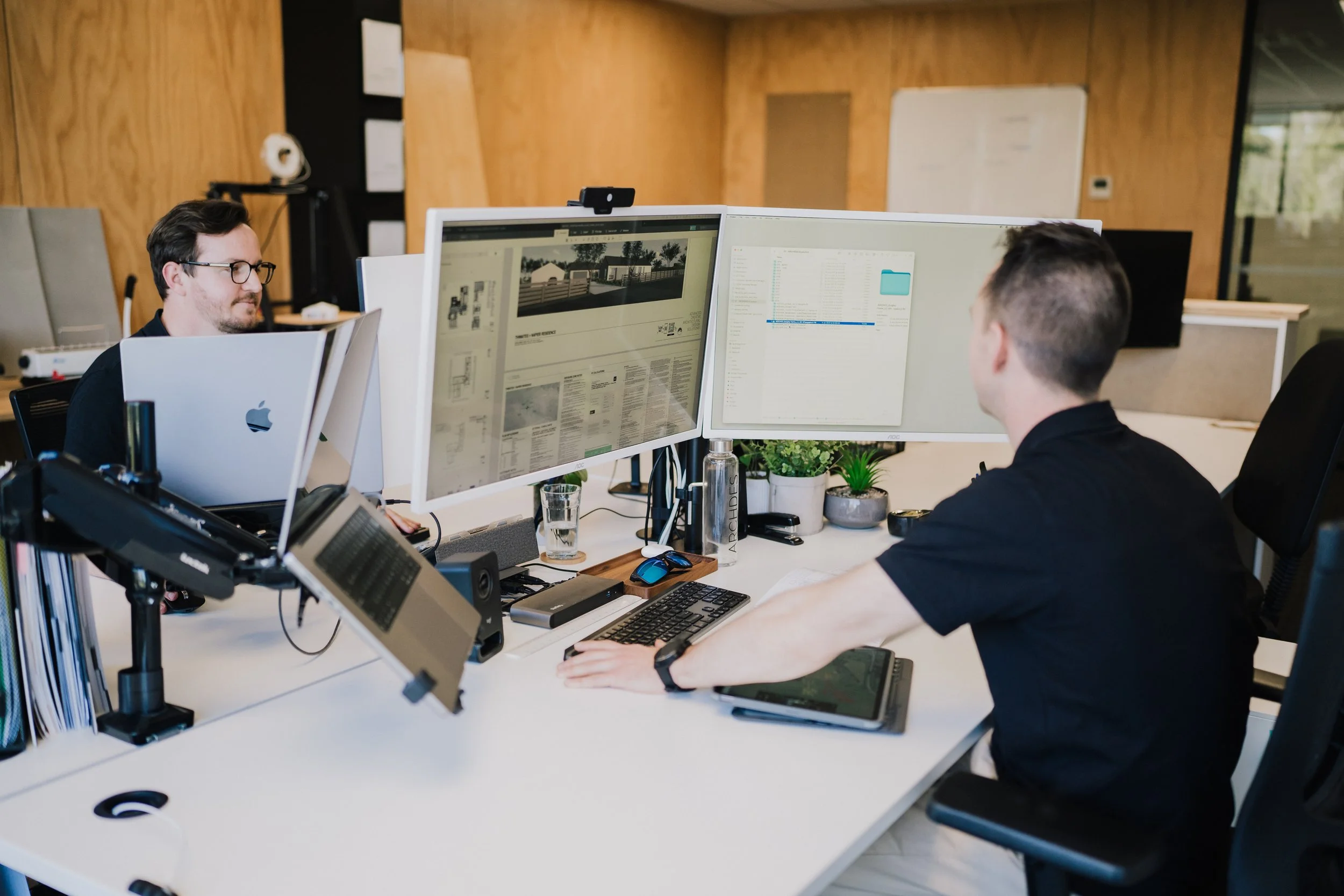 Two men working at a shared desk with dual computer monitors in an office with wooden walls and a glass wall in the background.