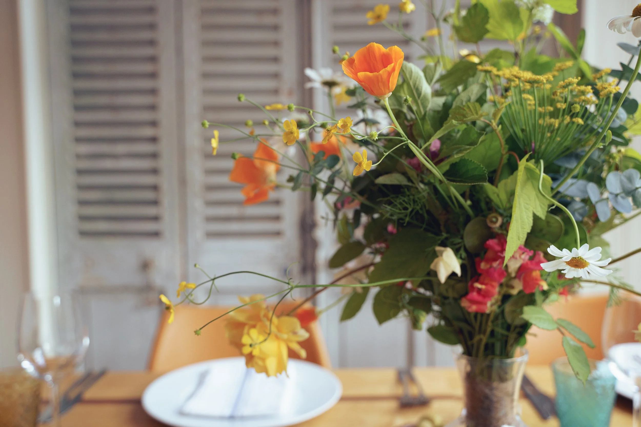 A large vase filled with a colorful assortment of flowers including orange, yellow, pink, white and purple blooms, on a wooden table set for a meal with plates and glasses, with a wooden louvered door in the background.