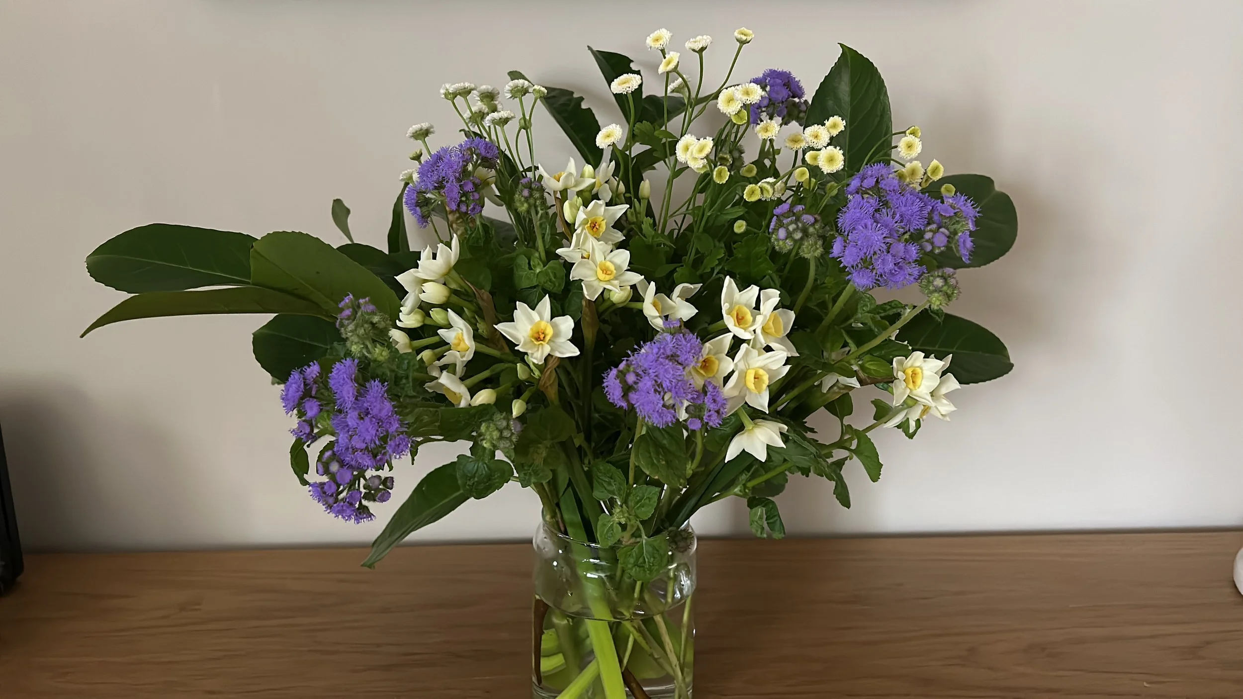 A glass vase filled with a colorful bouquet of flowers, including purple, white, and yellow blooms, placed on a wooden surface against a plain wall.