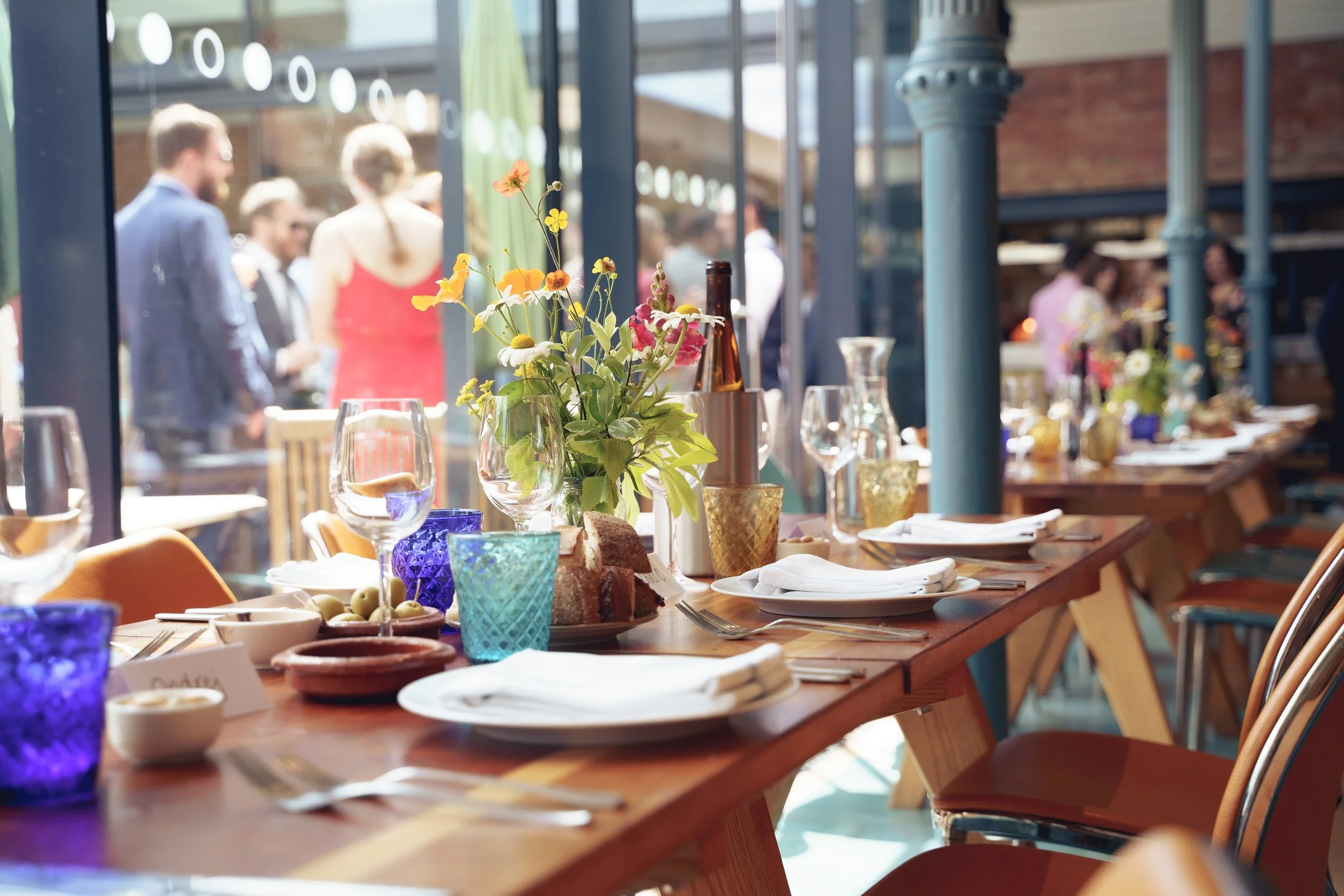 A decorated dining table set for a meal with plates, glasses, colorful cups, and a floral centerpiece, with people mingling outside through large glass windows.