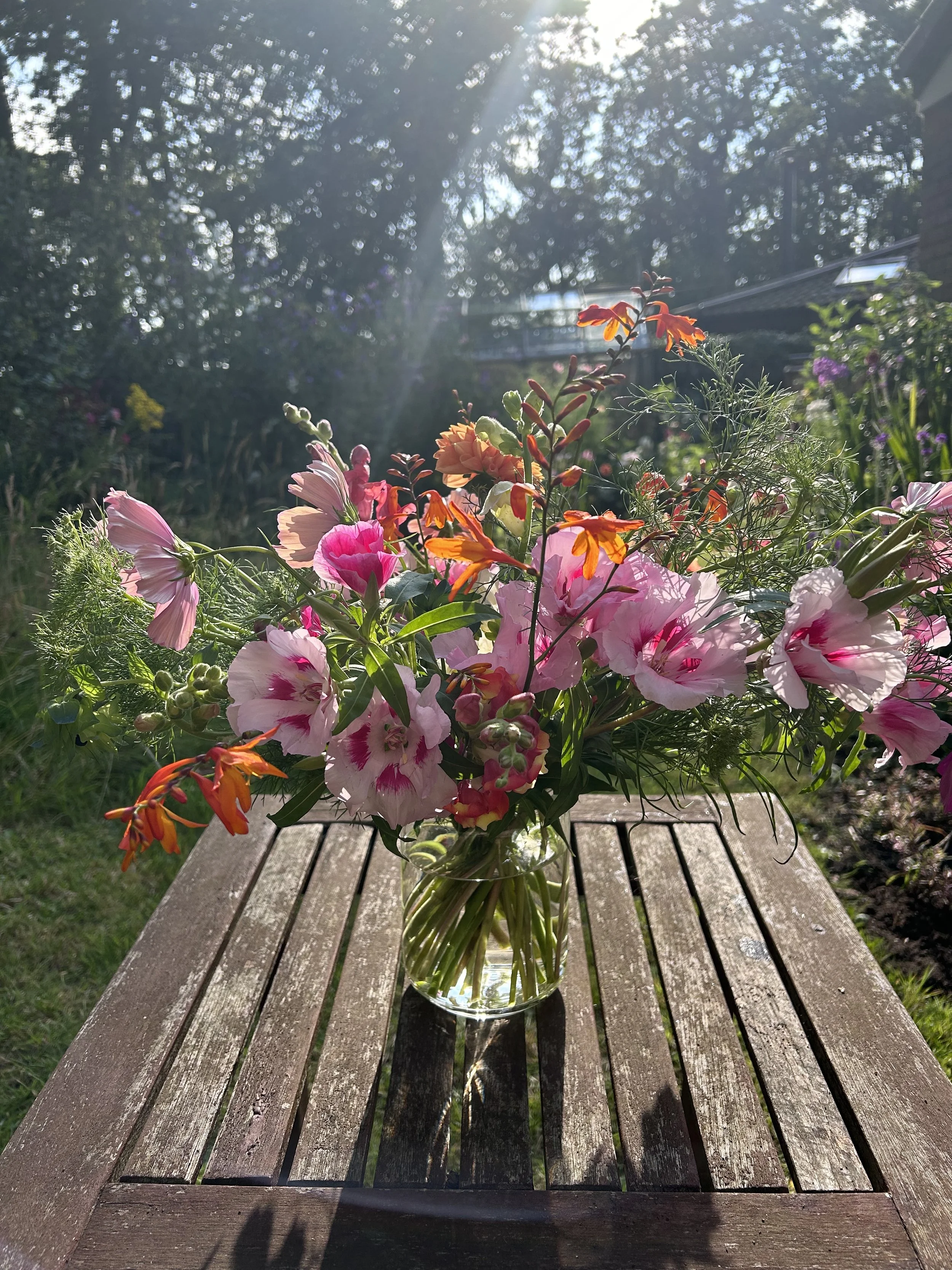A glass vase filled with a colorful bouquet of pink, orange, and peach flowers sitting on a wooden outdoor table with sunlight shining through the trees in the background.