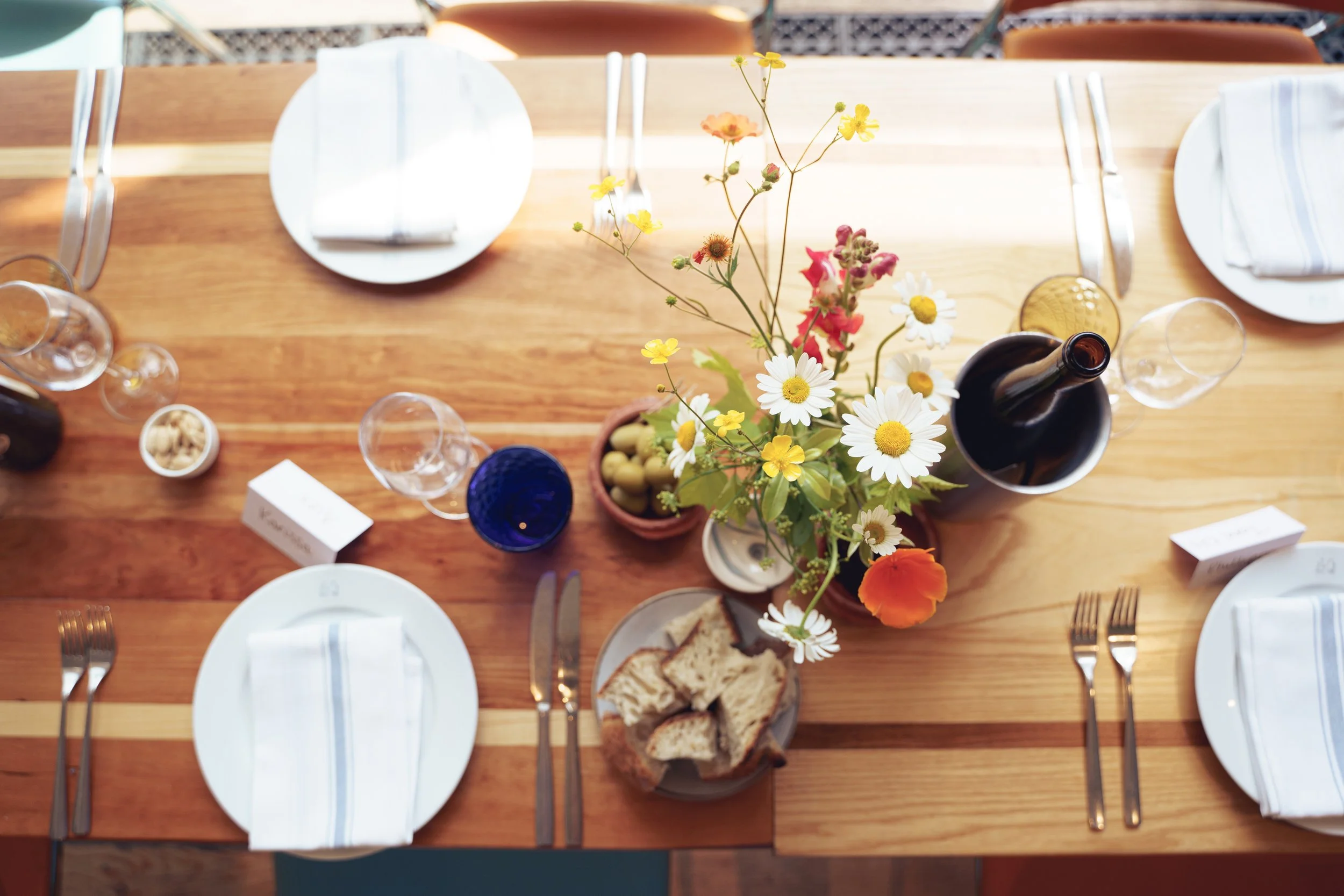 A wooden dining table set for a meal with four white plates, silverware, glasses, a small bowl of peanuts, a vase with colorful flowers, a bottle of wine in an ice bucket, a bowl of bread, and place cards.