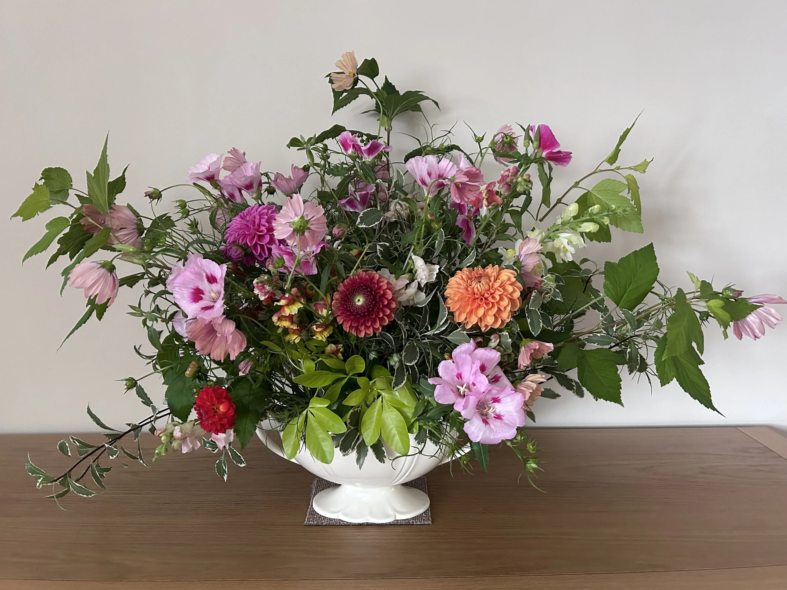 A floral arrangement in a white vase with a mix of pink, red, white, and orange flowers and green leaves, placed on a wooden surface against a plain white background.