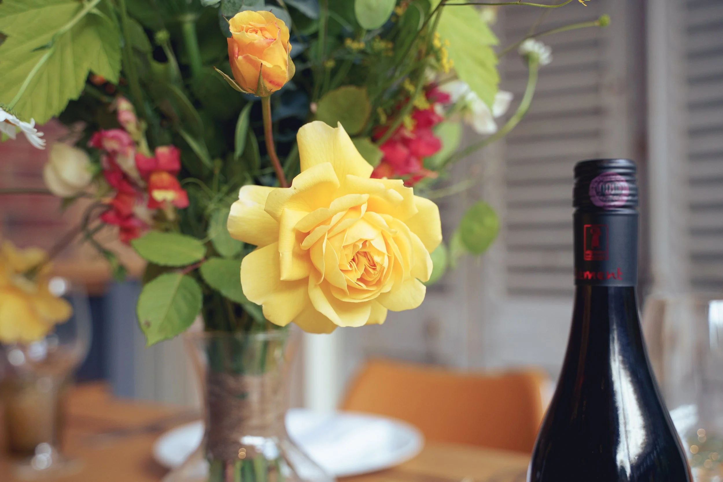 A close-up of a yellow garden rose in a vase with other seasonal mixed flowers in the background, and a wine bottle in the foreground.