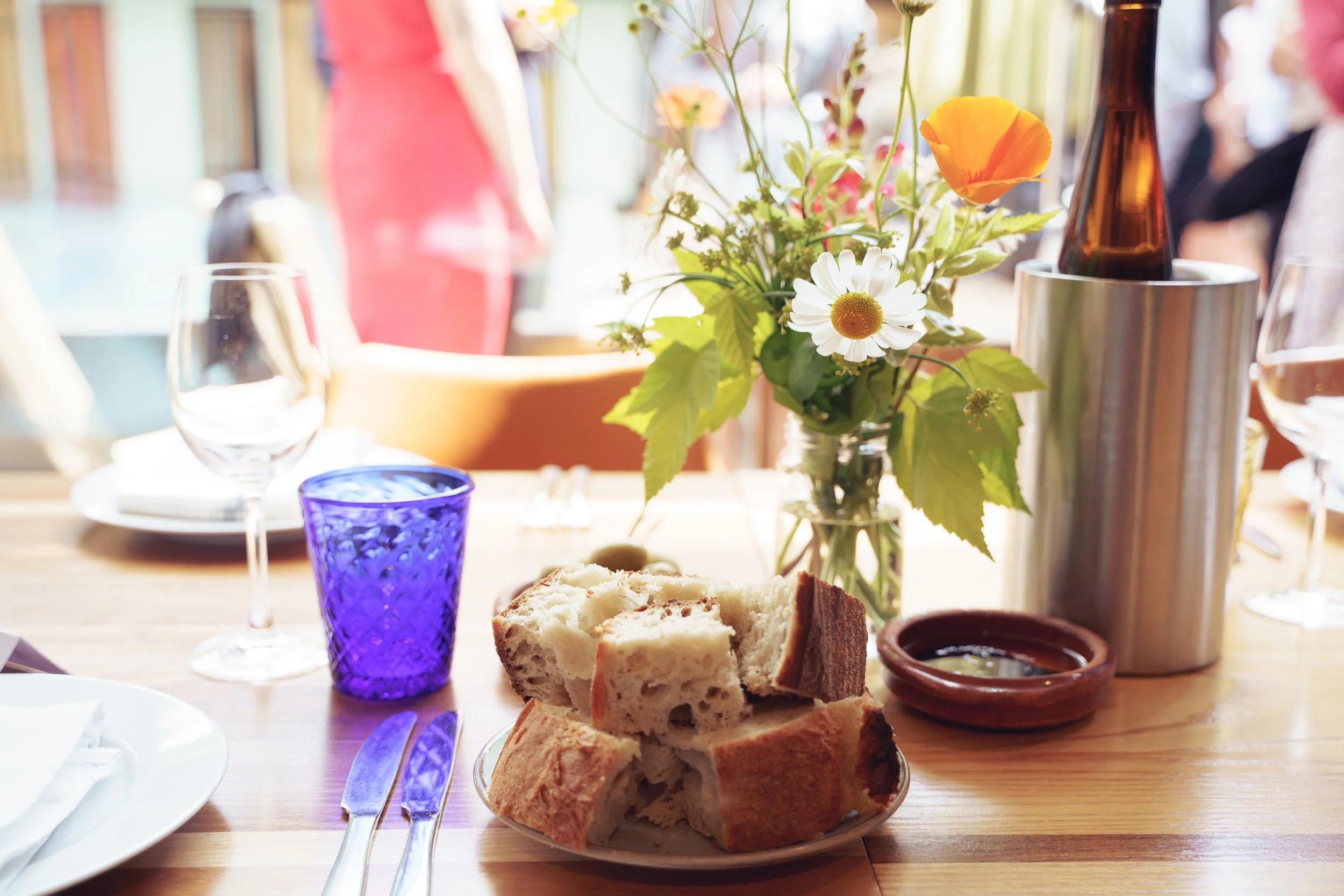 A table set for a meal with a loaf of bread, a glass of blue-tinted water, an empty wine glass, a small bowl of olive oil, a vase of mixed flowers, and a metal ice bucket, with other tables and people in the background.