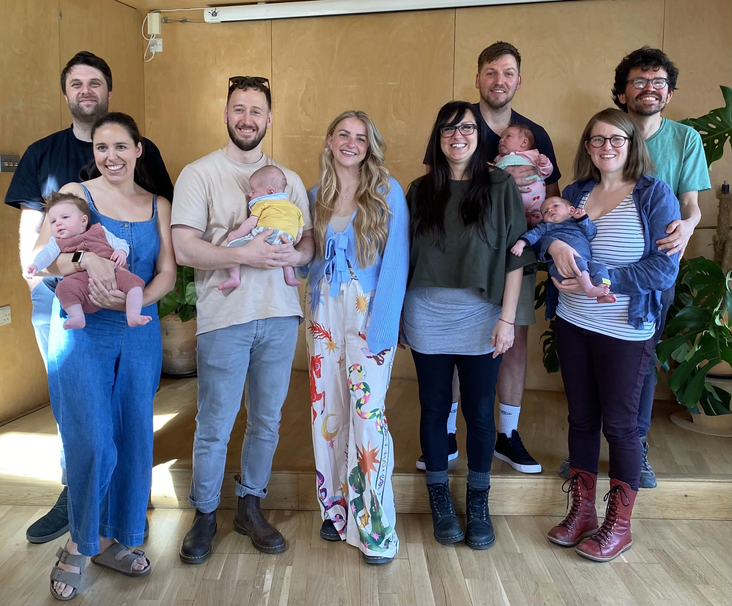 A group of ten adults, including four women and six men, and five babies, standing in a room with wooden walls and potted plants, smiling for the photo.