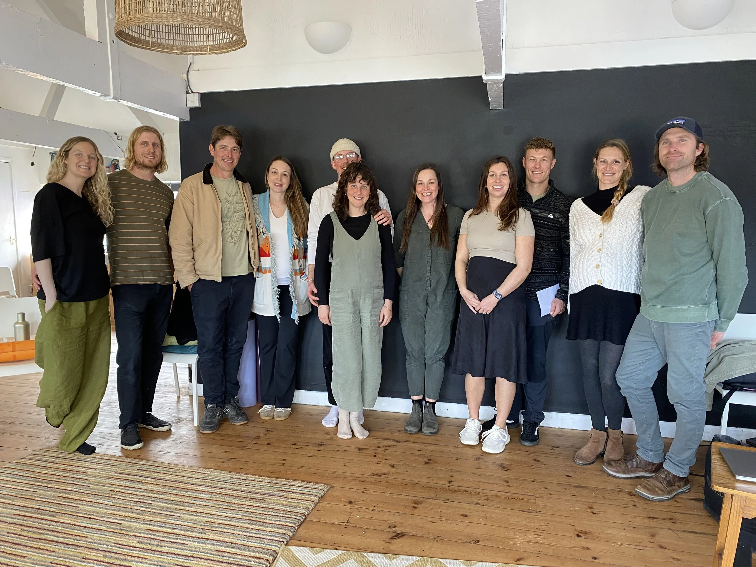 Group of twelve people standing together indoors, smiling, against a dark wall.