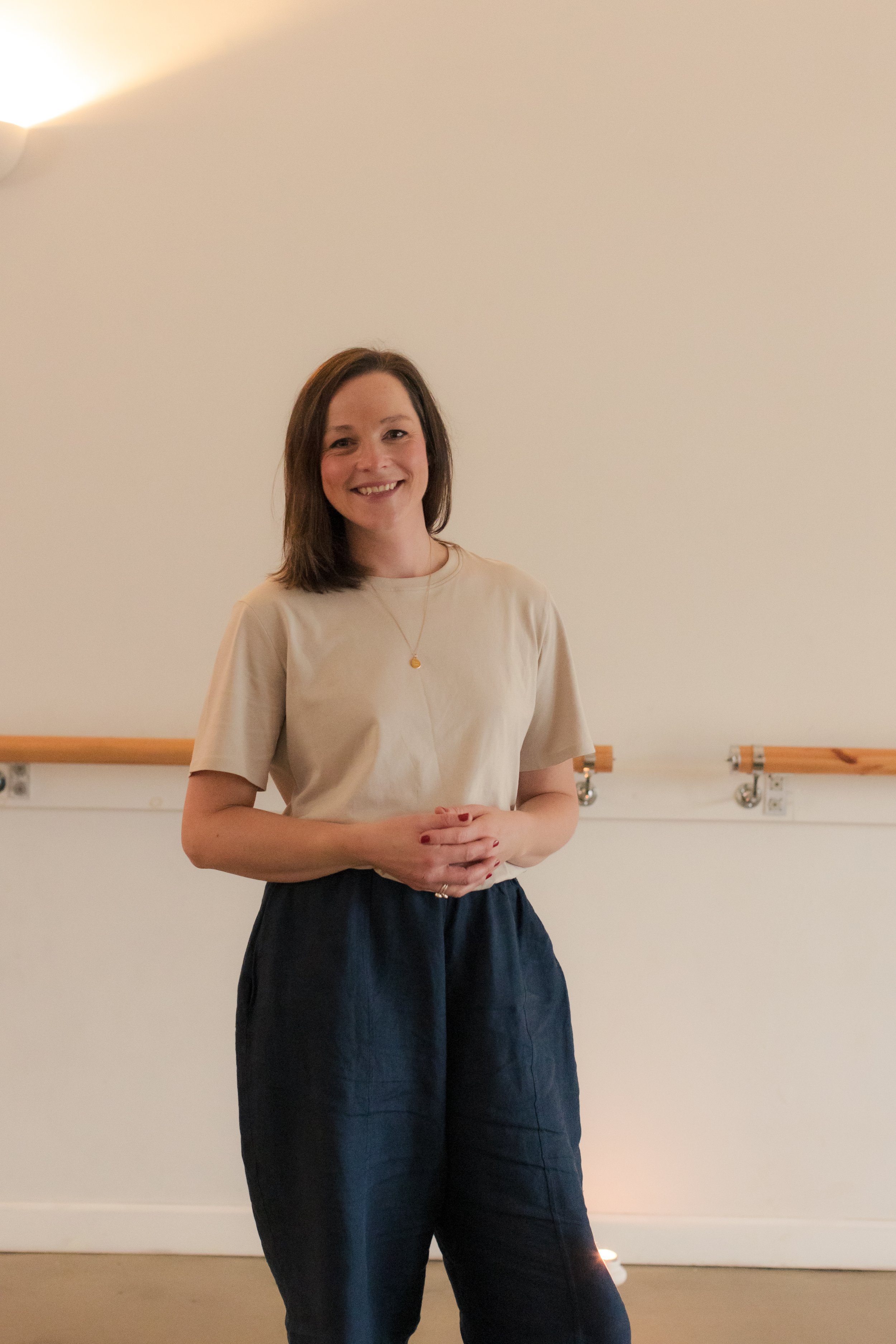 A woman with brown hair, wearing a beige T-shirt and dark pants, standing and smiling in front of a plain wall with a wooden handrail.