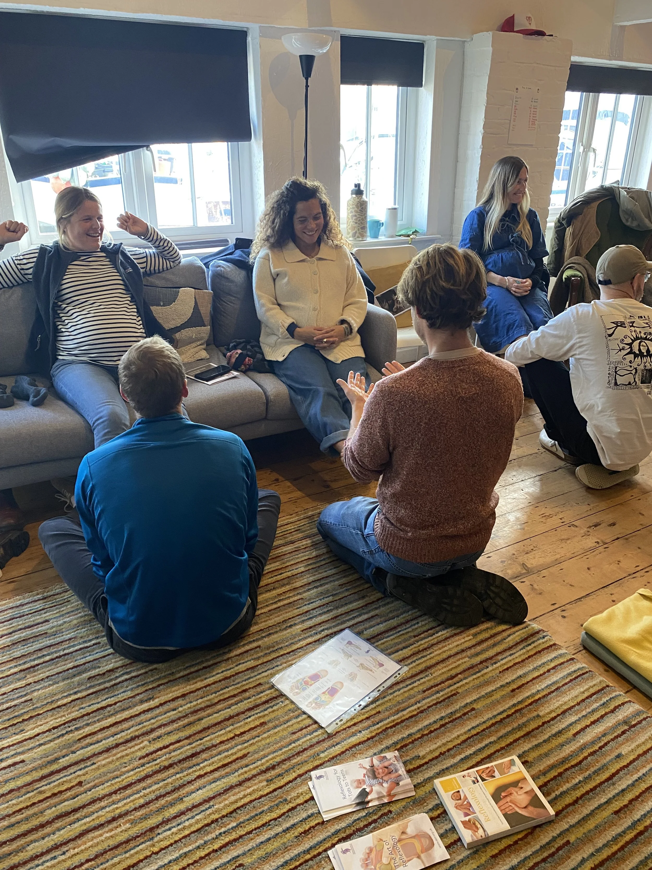Group of people engaging in a discussion in a cozy living room with wooden floors, a striped rug, and large windows, some sitting on a sofa and others kneeling on the floor.