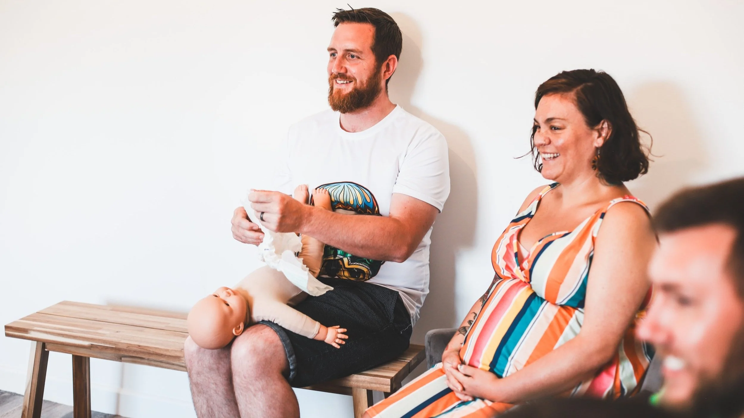 Man and woman sitting and smiling during a gathering or class, with a baby doll on the man's lap, in a room with white walls.