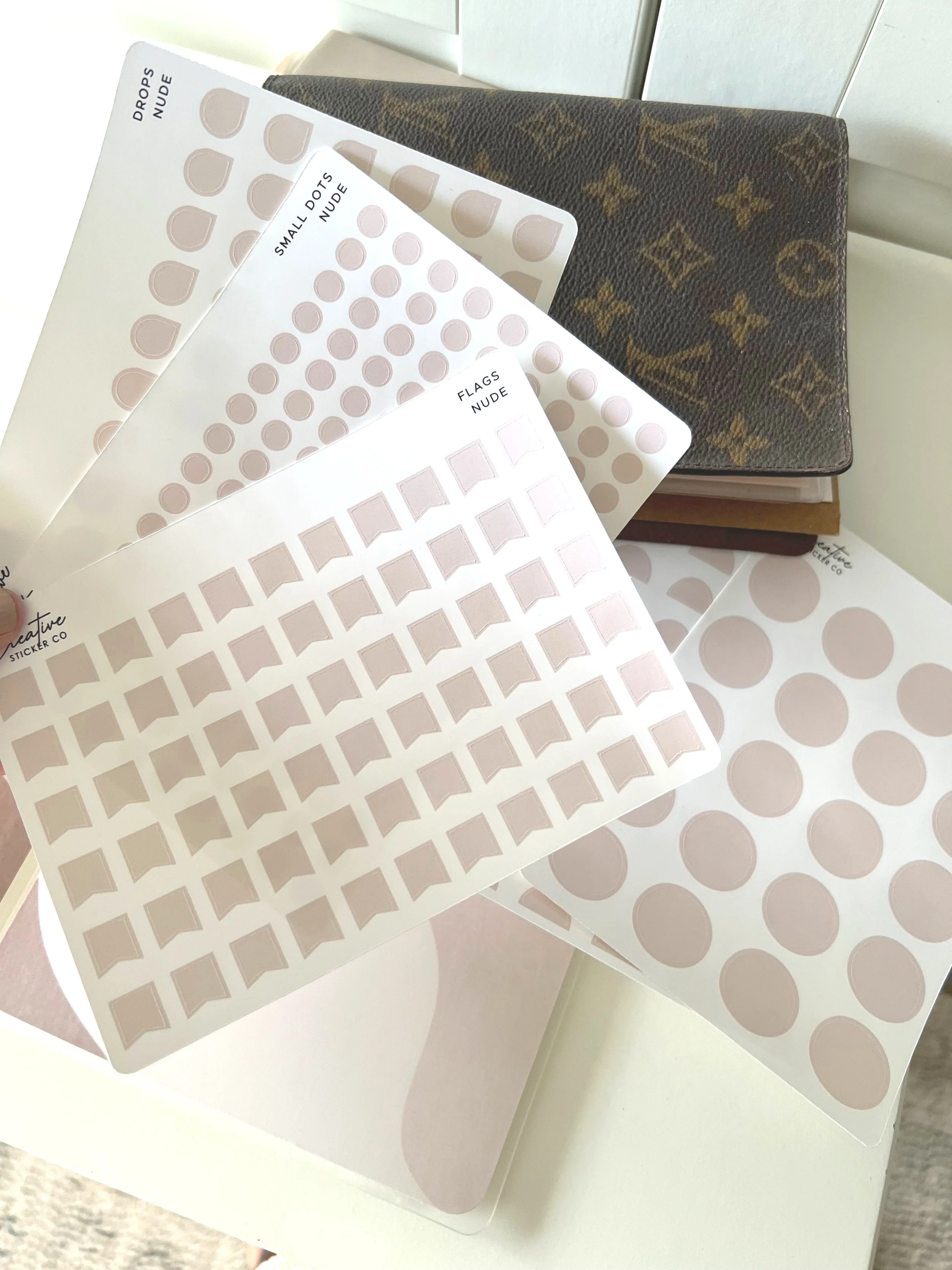 A collection of blank sticker sheets in various shapes and sizes, including drops, small dots, and flags, held in front of a Louis Vuitton wallet and a stack of books.