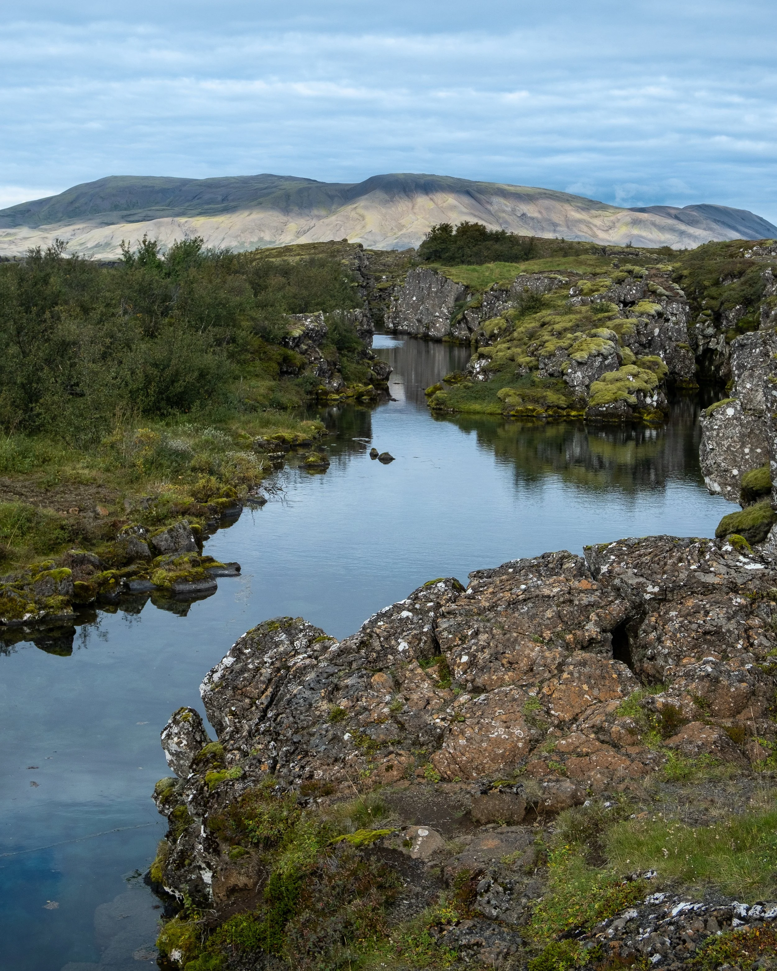 A river flowing through a rocky and mossy landscape with hills in the background under a blue sky with clouds.