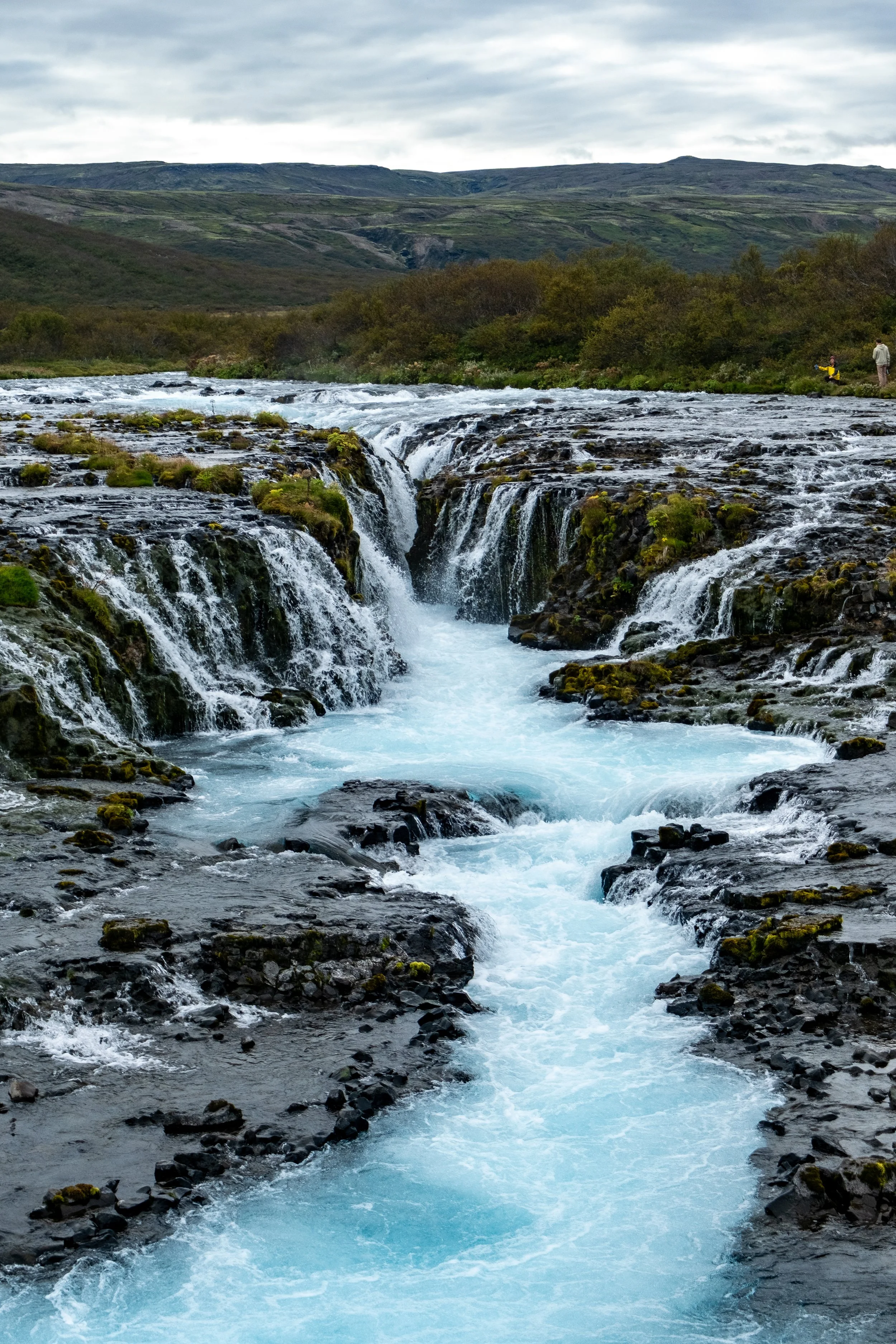 A scenic landscape of a river with small waterfalls, surrounded by green bushes and hills in the background, under a cloudy sky.